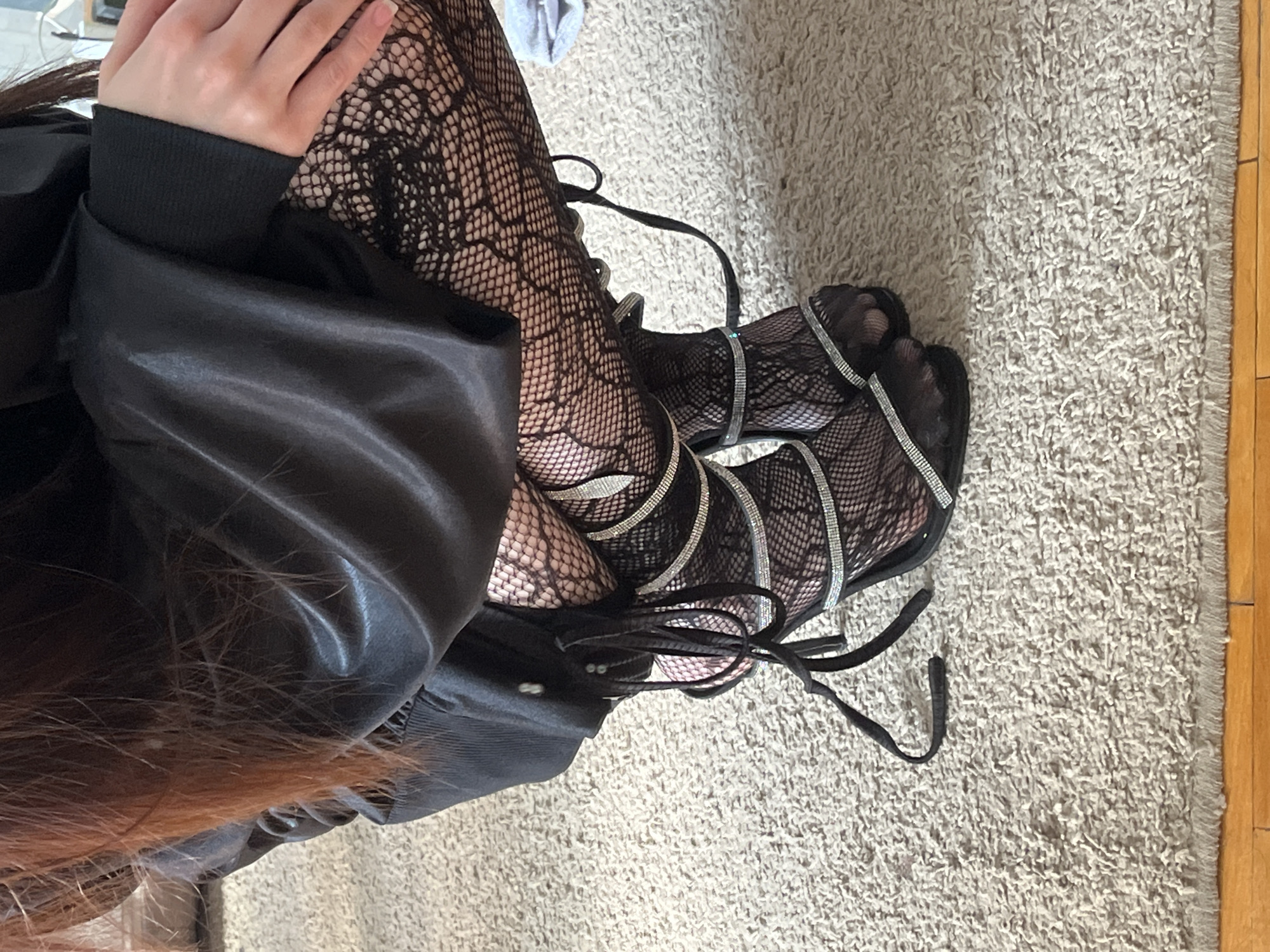 An overhead, close-up shot captures a person seated or kneeling on a light-colored, shaggy rug, showcasing their legs and footwear in an indoor setting. The scene is located in Ковалёво, Russia.
The subject's lower body is central to the image. They are wearing black, intricate fishnet stockings with a distinct, web-like pattern. On their feet are black, open-toed high-heeled sandals featuring multiple thin straps, heavily embellished with sparkling silver rhinestones or glitter that wrap around the ankles and lower legs. Black laces or ties from the sandals are visible on the rug.
A black garment, appearing to be made of a smooth, possibly satin-like or faux leather material, covers part of their torso and legs. A black ribbed cuff is visible on a sleeve near a hand with neatly manicured nails. Long, dark brown hair with visible reddish-brown highlights cascades over the black clothing.
The background consists primarily of a textured, light beige or off-white shag carpet or rug. Along the far right edge of the frame, a strip of light brown wooden flooring is visible, indicating the boundary of the rug within an interior space. No discernible activity beyond posing is evident. There is no visible text in the image.