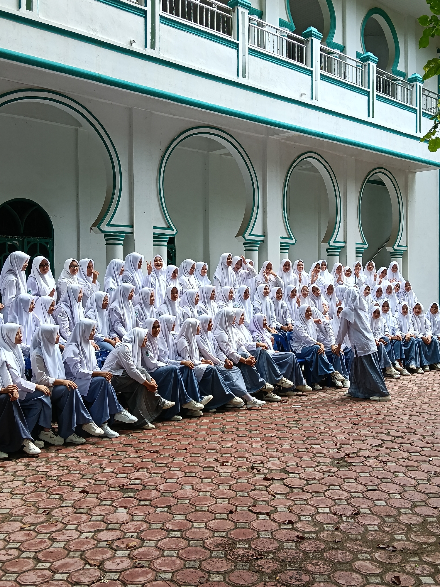 The image depicts a large group of young women, presumably students, gathered outdoors in Lhokseumawe, Indonesia. They are dressed in school uniforms consisting of white long-sleeved shirts, white hijabs, and either blue or grey skirts/trousers, largely paired with white sneakers. Most of the students are seated in multiple rows on chairs, arranged in a tiered formation, facing the camera with cheerful expressions. In the foreground on the right, one student stands, facing and seemingly addressing or interacting with the seated group.
The backdrop is a striking white building showcasing Islamic architectural elements, characterized by prominent teal-green decorative trim outlining arches, balconies, and structural features. The lower part of the building features several large arched recesses, while an upper level has a balcony with silver metal railings. The ground is paved with reddish-brown interlocking bricks, some of which are hexagonal or circular, with a few scattered leaves.
The activity appears to be a group gathering or photo session, marked by a lively and positive atmosphere. Many individuals are smiling, laughing, and making hand gestures like peace signs. The lighting is soft and natural, suggesting it is daytime, possibly morning or late afternoon. While some white text is faintly visible on a bag or clothing item worn by one of the seated students, it is too indistinct to be read. The scene conveys a sense of camaraderie and joy among the students.