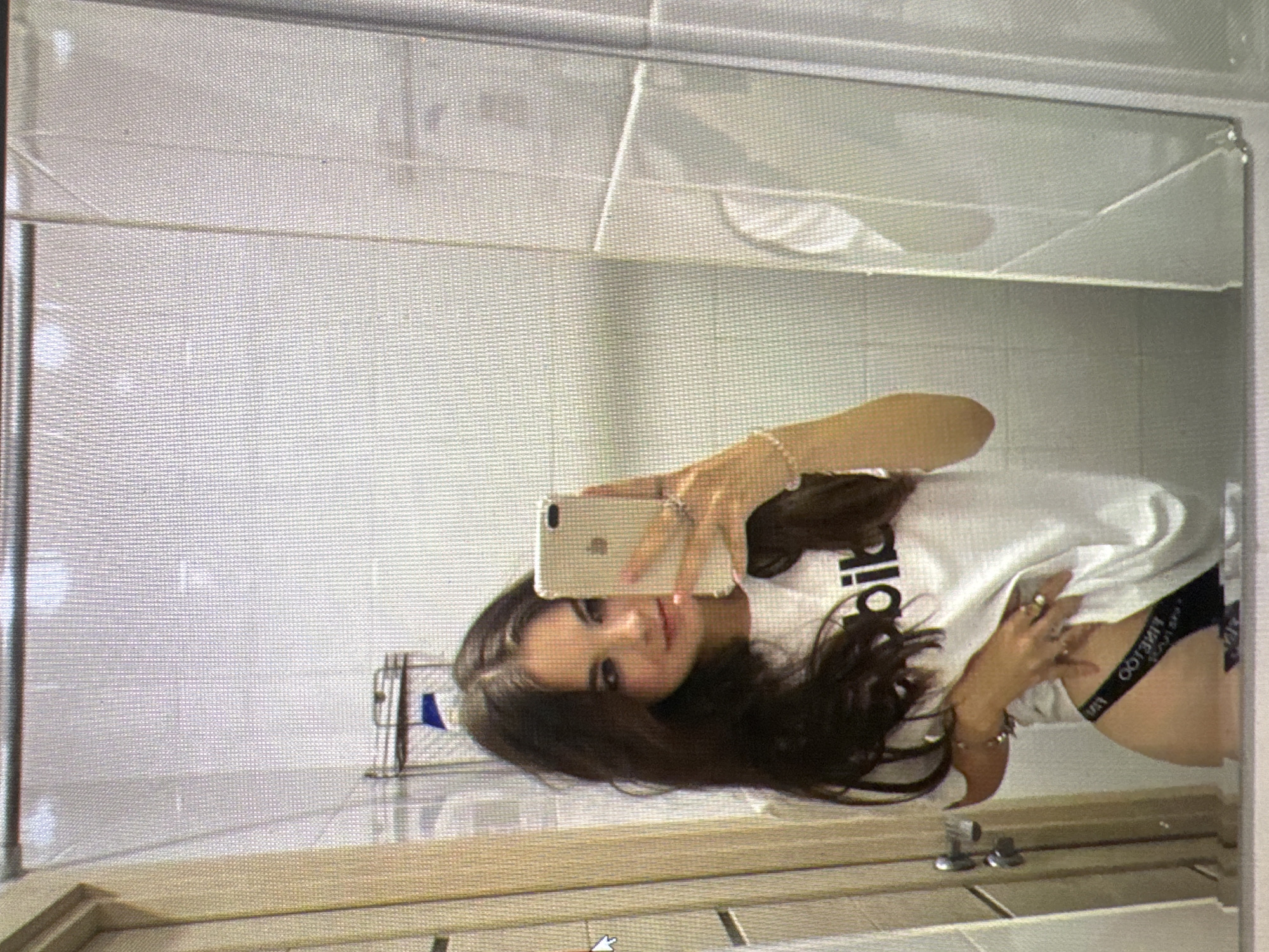 The image shows a young woman taking a mirror selfie. She is wearing a white t-shirt with black lettering that appears to say "adidas" or a similar logo, and black underwear with a visible band. She has long, dark hair and is wearing several rings on her fingers and a bracelet on her wrist. The setting appears to be a bathroom, with white tiled walls and a wooden countertop or vanity visible in the foreground. A mirror reflects the woman and the room. The lighting suggests it is daytime, possibly indoors with artificial light. The woman's expression is neutral. The image is oriented sideways.