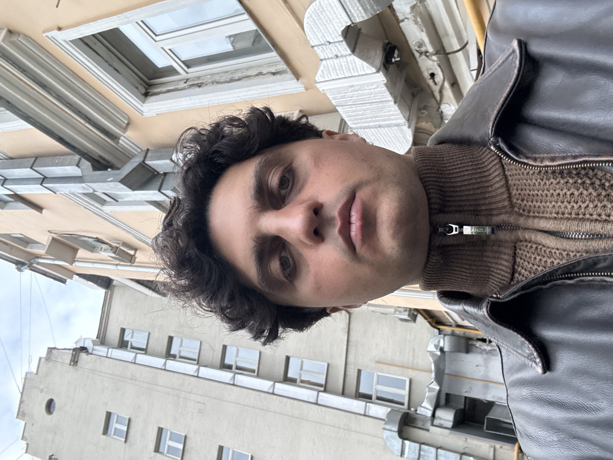 A man is taking a selfie in what appears to be Moscow, Russia. He is wearing a brown sweater and a dark leather jacket. The background features buildings with distinct architectural details. The sky is overcast. Windows and ventilation systems are visible on the facade of the buildings. The perspective suggests he's looking upward, possibly from a narrow street or courtyard.