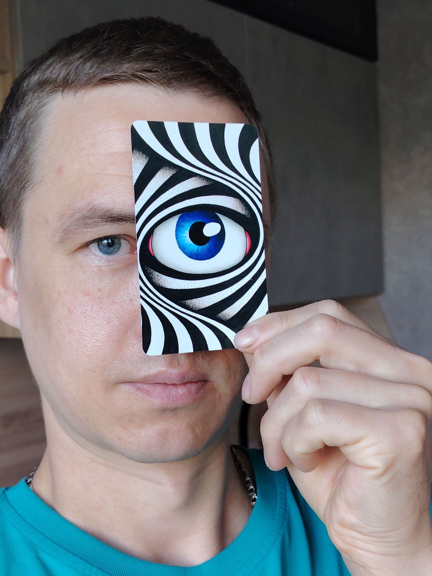 An adult male, presumably in Chaikovsky, Russia, is captured in a close-up, chest-up shot indoors, likely during the daytime. He is light-skinned with short, dark hair and a neutral expression. His left eye, which is blue, is fully visible. He is wearing a teal or turquoise t-shirt and a subtle chain necklace.
The central activity involves him holding a rectangular card vertically in front of his face with his right hand, covering his right eye. The card features a striking, surreal design: a large, vivid blue eye with a black pupil, surrounded by swirling black and white stripes that create an optical illusion effect, reminiscent of an artistic interpretation of an iris scanner or a hypnotic pattern. His fingers are partially visible as he grips the card.
The background is softly blurred, showing parts of a wall or interior structures, suggesting a domestic or simple indoor setting. There are no specific cues within the image itself to identify the location as Chaikovsky, Russia, beyond the provided context. No visible text is present in the image. The overall impression is a playful or artistic presentation of the optical illusion card.
