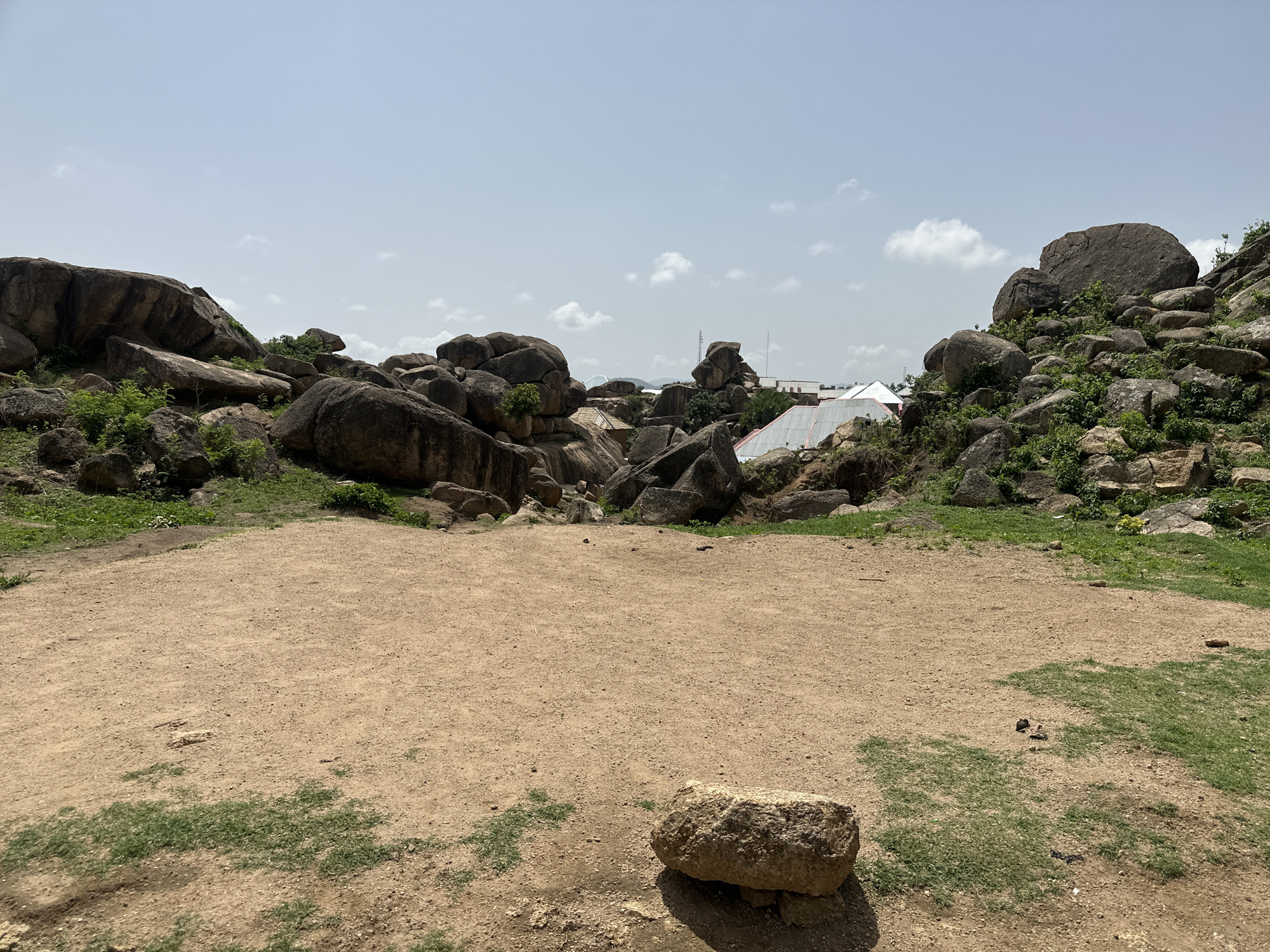 The image depicts a landscape with rocky formations and sparse vegetation, likely located in Jos, Nigeria, based on visual cues. In the foreground, there is a predominantly sandy area with scattered patches of grass. A large rock rests on the lower portion of this area. The middle ground features an arrangement of large, dark-colored rocks and boulders, interspersed with greenery. In the distance, a structure with a white roof is visible. The sky is overcast with a few clouds. No people are visible in the image.