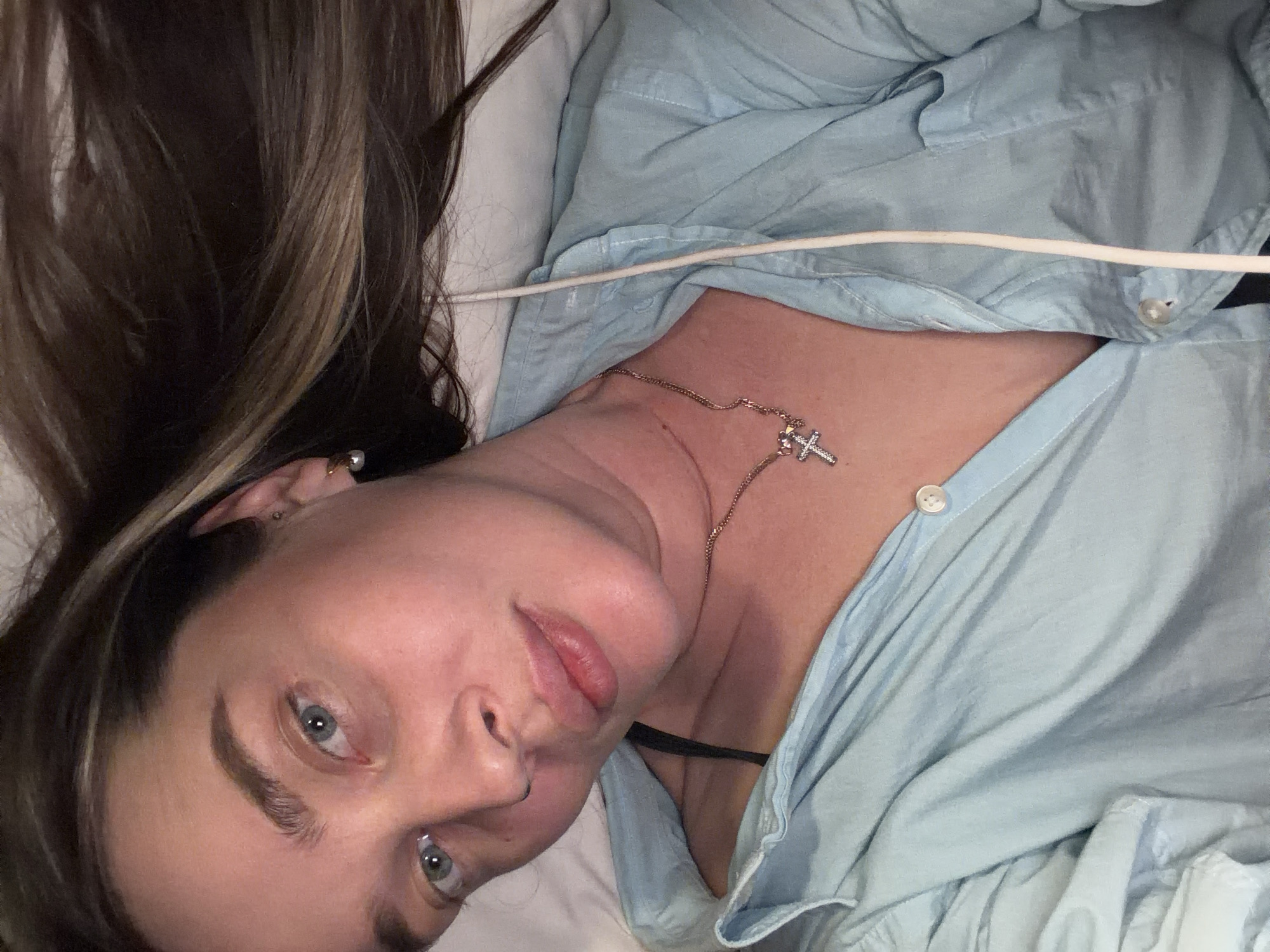 A woman lies on a white pillow wearing a light blue button-down shirt and a gold necklace with a cross pendant. Her hair is dark with light highlights. She has fair skin and blue eyes. A white wire or cord is visible across her shirt. The photo appears to be taken indoors with soft lighting. There is no indication of the location being Moscow, Russia.