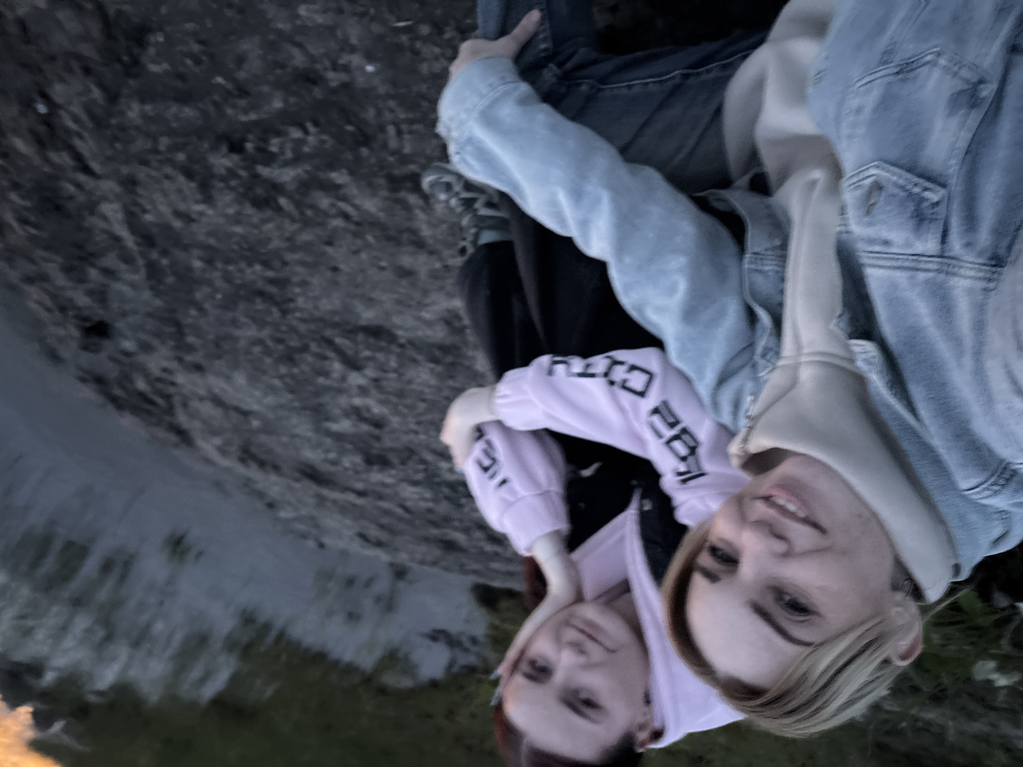 Two young women are pictured in a selfie taken outdoors during daylight. The woman in the foreground, on the left, has blonde hair and is wearing a denim jacket over a cream-colored turtleneck. She is smiling at the camera. To her right, a woman with red hair is leaning her head on her hand, looking off to the side. She is wearing a pink hoodie with black lettering. They are both sitting on a dark, rocky surface next to a body of water, which appears to be a lake or river. The background shows the water and some greenery. The overall lighting suggests it might be late afternoon or early evening. The scene is set in Shuiskaya Chupa, Russia, where the landscape features natural elements like water and rocky terrain, characteristic of the region's natural beauty.