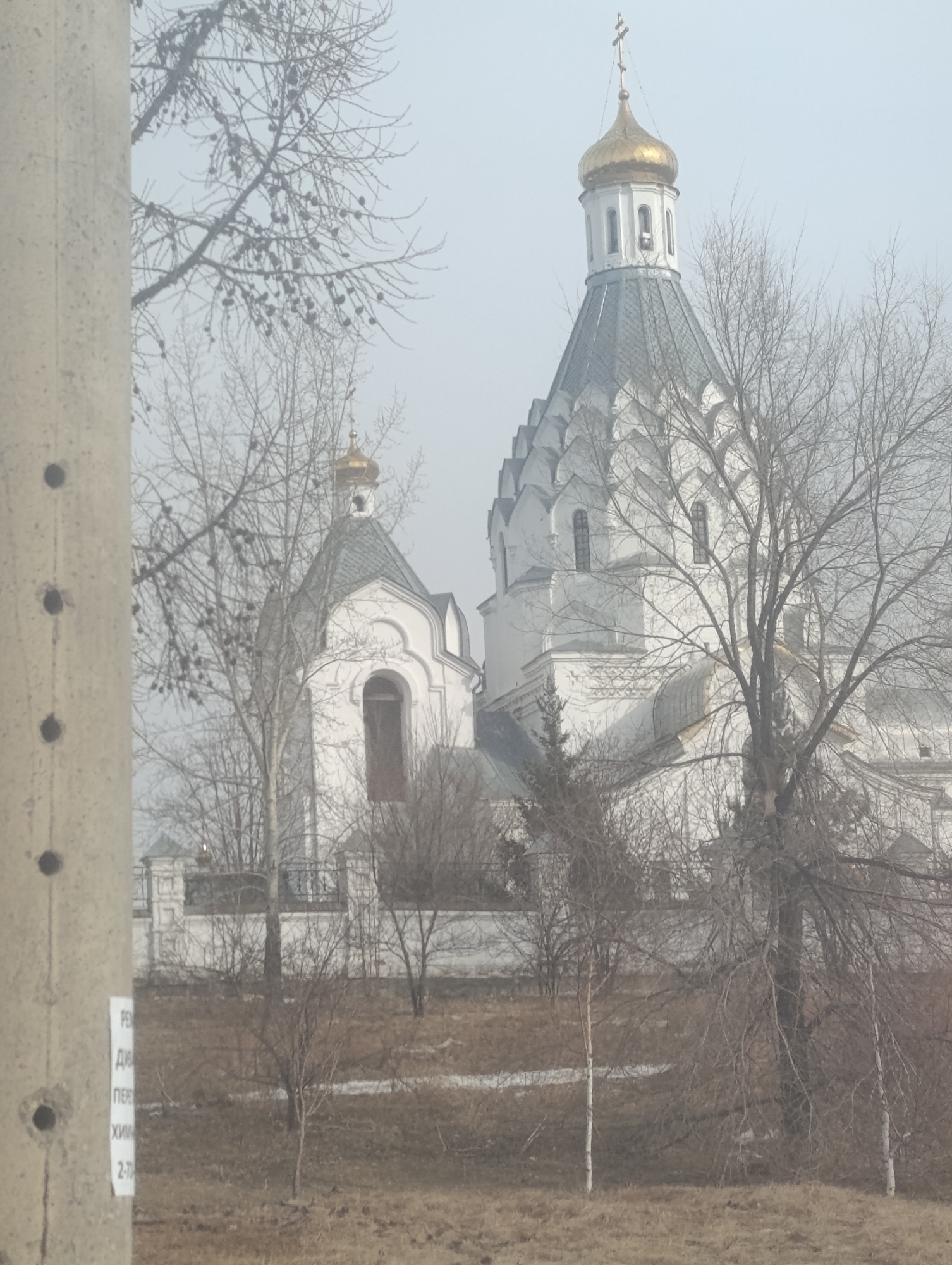 The image shows a white Russian Orthodox church with a golden dome, partially obscured by leafless trees. The church is surrounded by a low wall and appears to be located in a grassy area. In the foreground, a concrete utility pole with faded Russian text is visible. The sky is overcast, creating a hazy, muted atmosphere. Based on the architectural style of the church, this may be located in Krasnoyarsk, Russia.