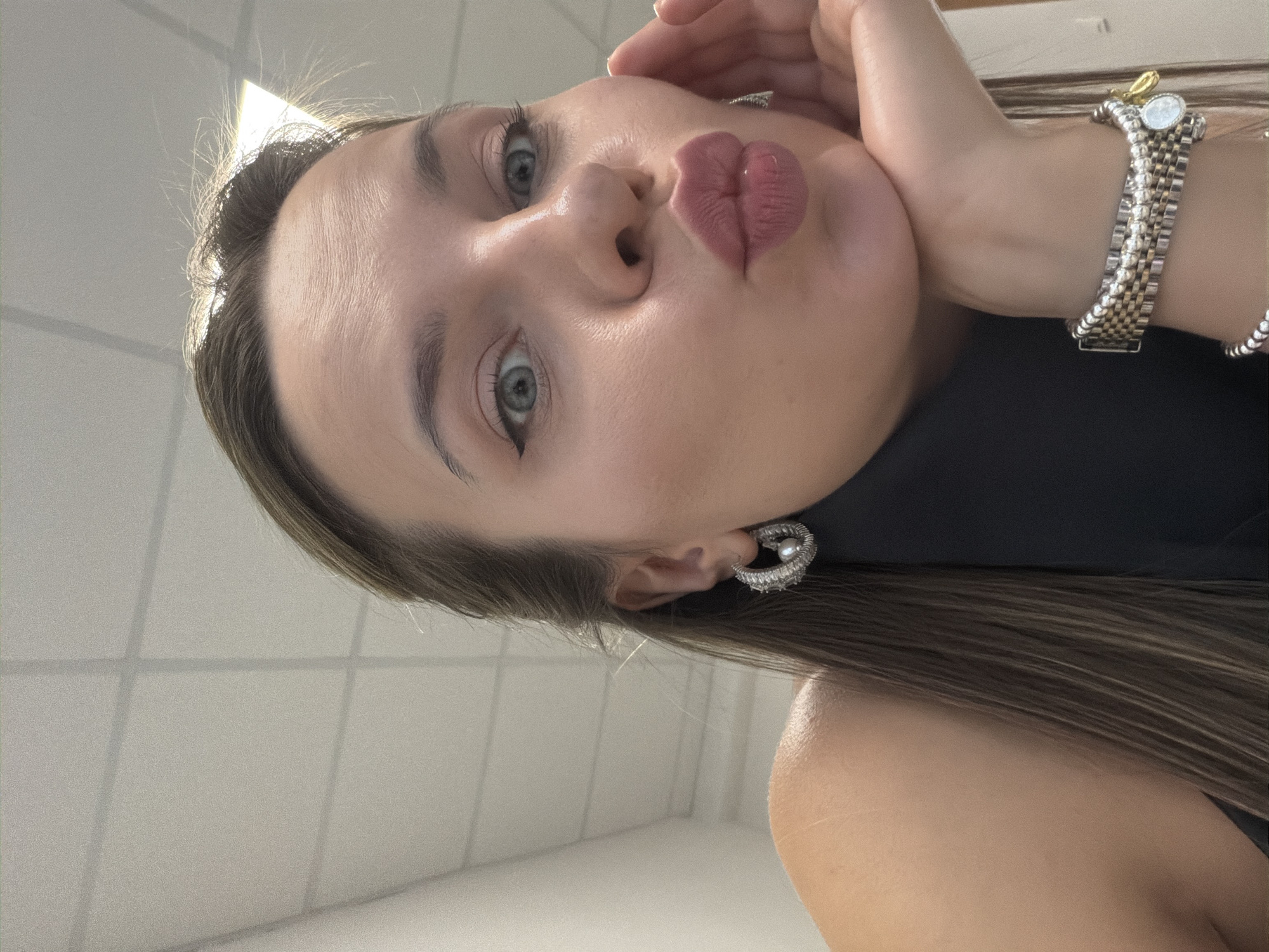 A young woman with brown hair and blue eyes, wearing a black turtleneck and silver hoop earrings, is looking at the camera with her lips pursed in a kiss. She is resting her chin on her hand. She is also wearing a silver bracelet with charms. The background appears to be an office with a tiled ceiling and fluorescent lighting. The image does not contain any information about the city of Homyel, Belarus.