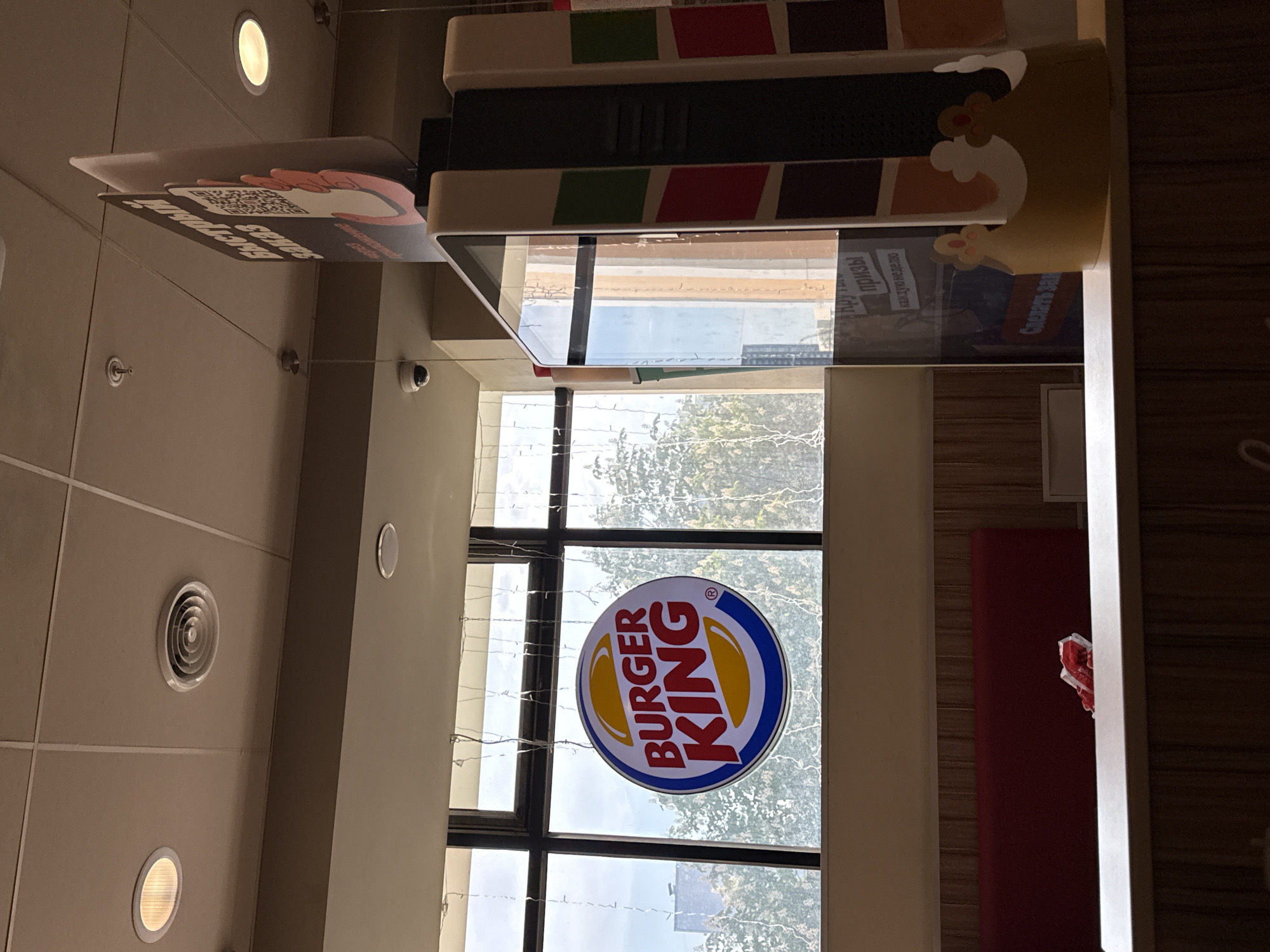 The photograph shows the interior of a Burger King restaurant. Visible is a window with the Burger King logo. A digital menu board hangs above, and the ceiling has multiple circular lights. The presence of text in Russian on the menu board implies a location in Russia, and Moscow is a possible location. There are no people visible in the image. The scene is lit by both artificial lighting and natural light coming through the window. A red booth seat is partially visible on the right side of the image.