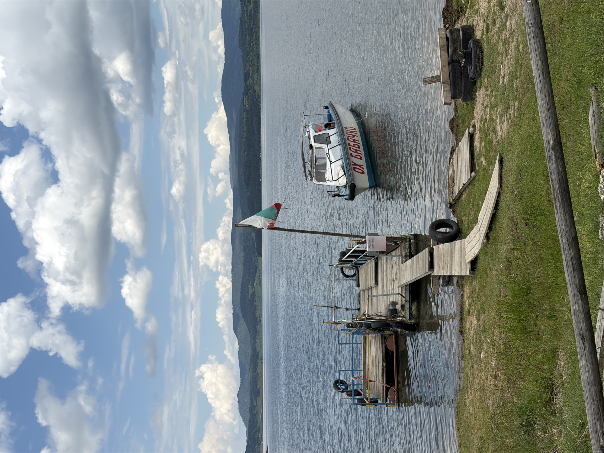 The image depicts a tranquil lakeside scene in Цигов чарк, Bulgaria, on a day characterized by a blue sky with numerous large, white cumulus clouds. In the background, a range of dark green, forested mountains stretches across the horizon, meeting a calm, grey-blue body of water, likely Batak Lake.
In the midground, a rustic wooden dock extends into the water from the grassy shore. The dock consists of multiple interconnected floating wooden platforms, equipped with rudimentary metal railings. Several old tires are integrated into the dock structure, serving as fenders or flotation. A wooden pole on the main dock section proudly flies a Bulgarian flag, displaying horizontal white, green, and red stripes. A small pink sign on the dock bears Cyrillic text: "РАЗХОДКИ (088 850 70)", which translates to "WALKS (088 850 70)", indicating boat excursions are offered, along with a contact number.
To the right of the dock, a white motorboat with a blue stripe on its hull is moored in the water. The boat has a small cabin, and red Cyrillic lettering on its side reads "ОХ БАБАЧКО". A small Bulgarian flag is also visible on the boat. A faint figure, possibly a child, can be seen inside the boat's cabin through a window.
The foreground consists of a grassy bank leading to the water's edge. Multiple wooden plank walkways connect the shore to the floating dock. On the grassy area to the right of the dock, a stack of old tires is present, along with a single tire lying flat. A rough, weathered wooden log or fence runs horizontally across the very bottom of the frame, partially obscured by grass. The water surface shows minor ripples, reflecting the sky and distant features.