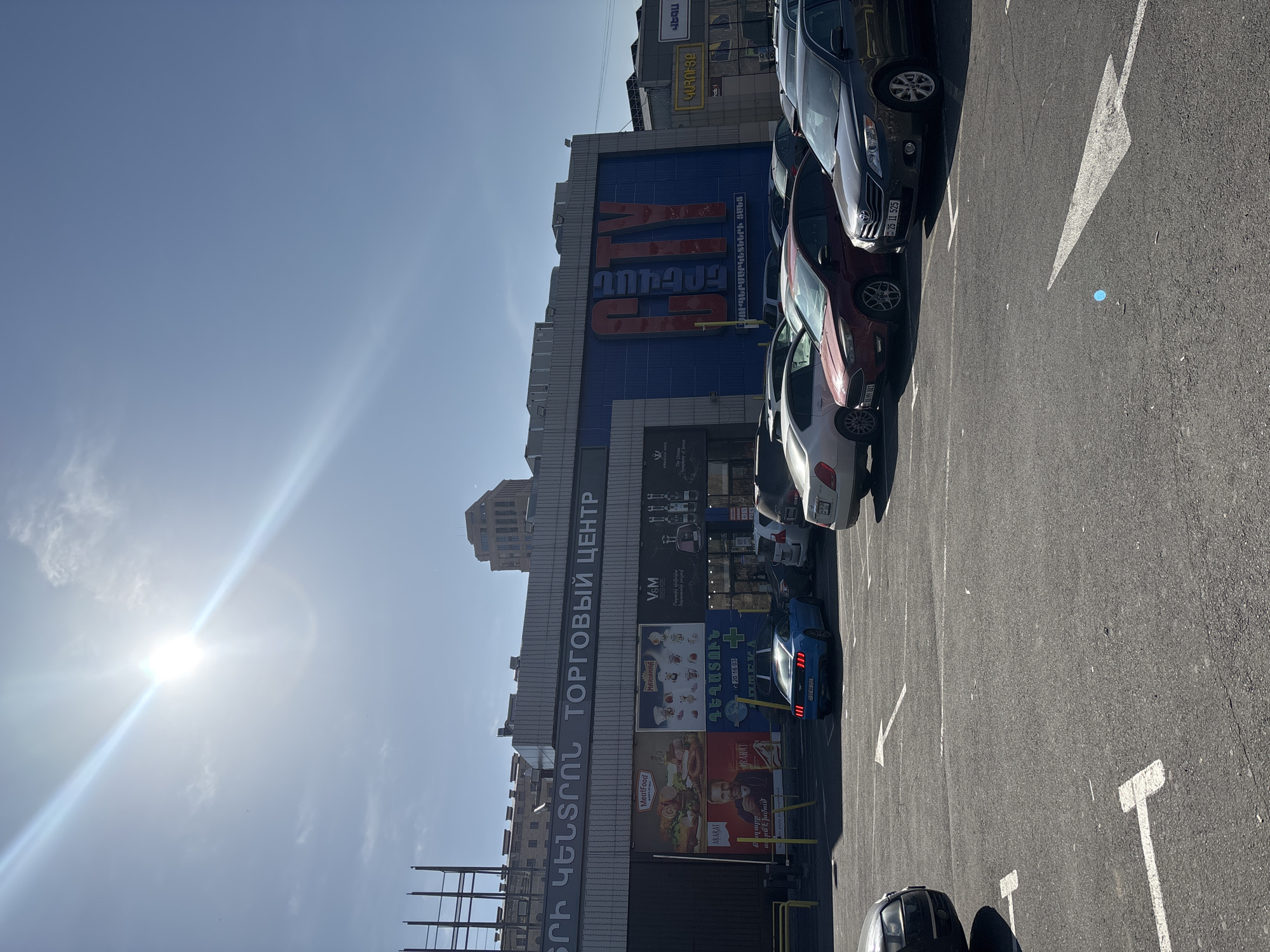 This is an outdoor shot of a commercial area in Yerevan, Armenia. The image is rotated counter-clockwise. Prominently visible is the "Erevan City" supermarket building, which has the name written in Armenian and Russian ("ԵՐԻ ԿԵՆՏՐՈՆ ТОРГОВЫЙ ЦЕНТР"). Adjacent to the building is a large parking lot with cars parked in it. On the building, there are advertisements and signage for different brands, including "V&M," "Marianne," "MeatFood," and "ARARAT." The sky is clear and bright, with a visible sun glare.