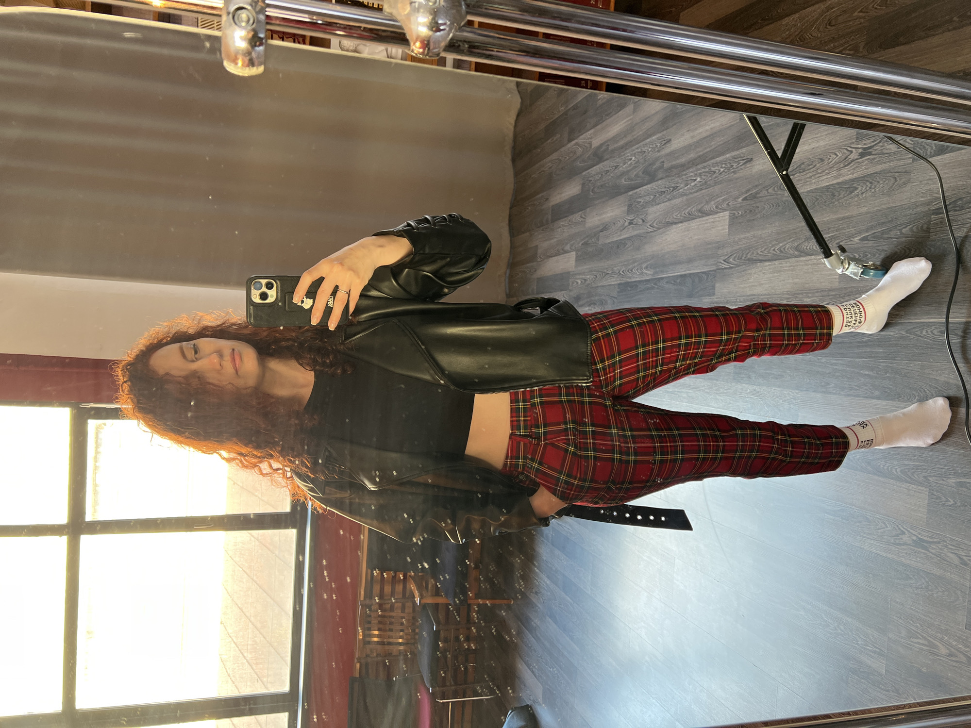 A young woman is taking a selfie in a mirror, likely indoors, with sunlight streaming in through a large window. She has long, curly red hair and is wearing a black crop top, red plaid pants, a black leather jacket, and white socks. She is holding a black smartphone in her right hand to capture the image. The mirror's frame is visible, with part of its support structure visible in the reflection. The floor is gray with a wood-grain pattern. The location cannot be confirmed as Saint Petersburg, Russia based on visual information.