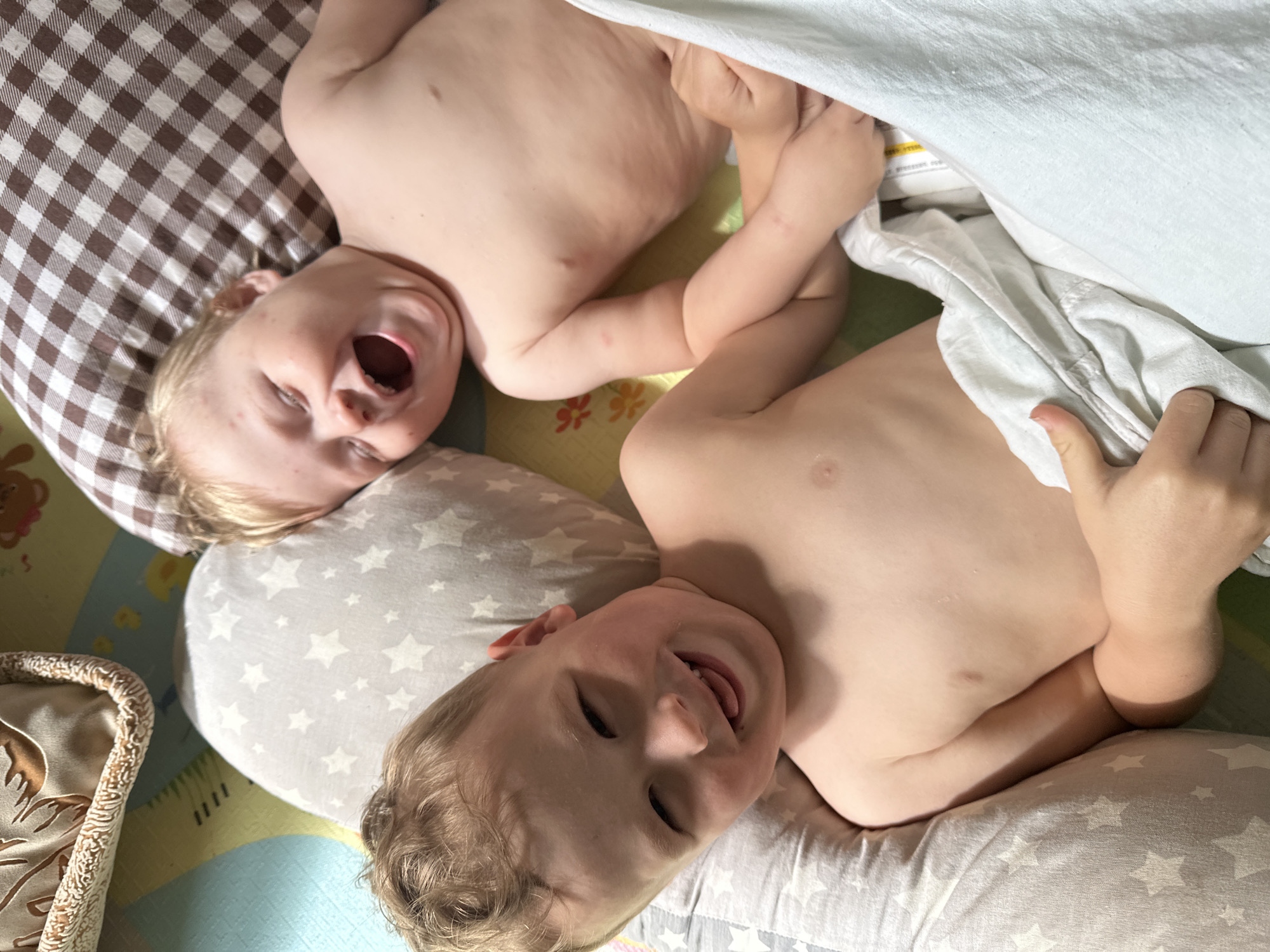 Two young children are lying on a patterned surface, possibly a play mat or bedding. The child at the top left is lying on a brown and white checkered pillow, mouth open in laughter. The other child is on a grey pillow with white stars, also smiling. The children are bare-chested and holding hands. The background is a combination of different patterns and textures.