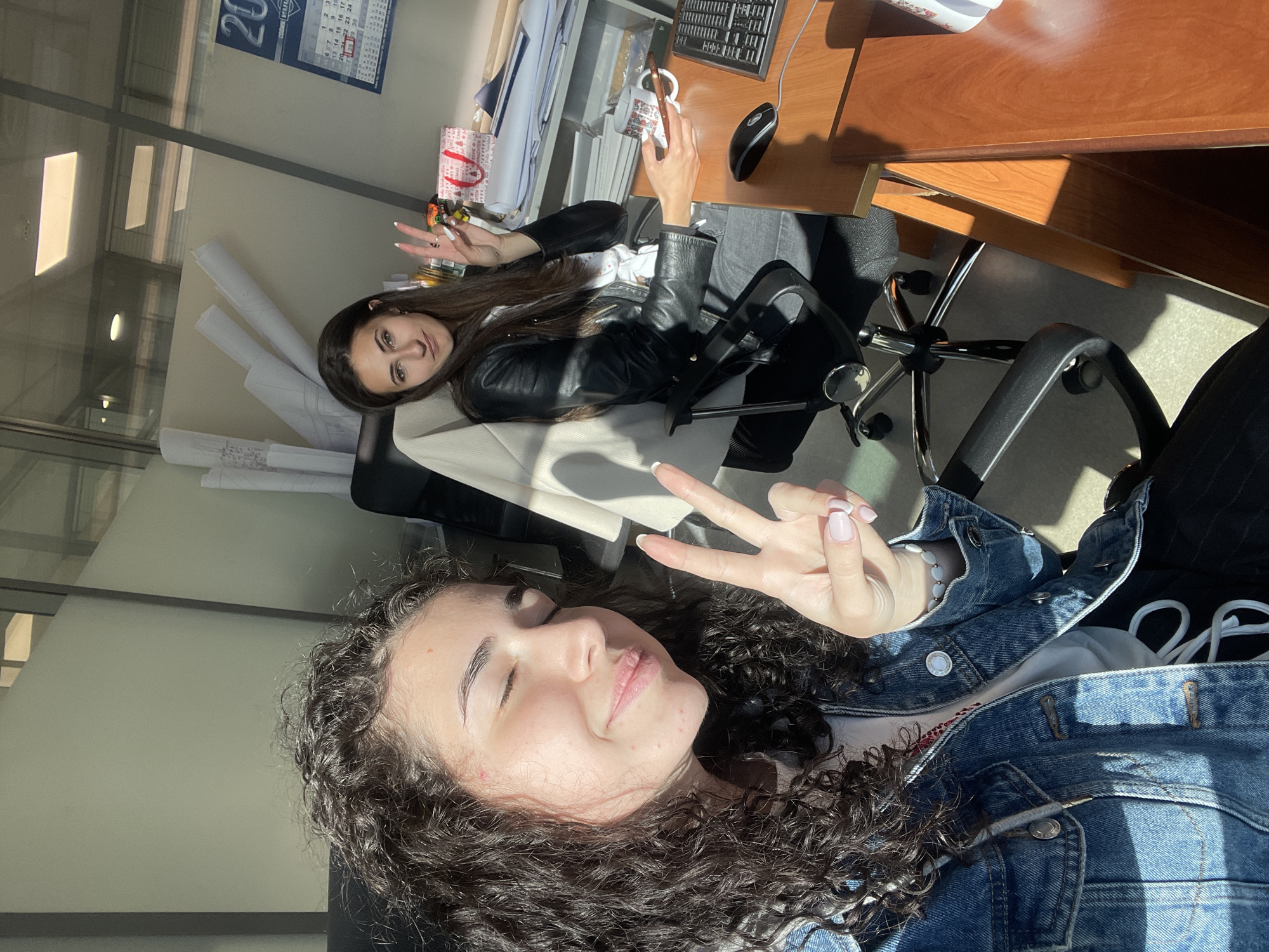 This media file depicts two young women posing happily in an office or studio setting in Sofia, Bulgaria. The image, likely a selfie taken from a low angle, is brightly lit by natural sunlight, casting strong shadows and suggesting a sunny daytime.
In the foreground, a woman with dark, curly hair, wearing a denim jacket, smiles with her eyes partially closed while making a peace sign with her right hand. Behind her, another woman with long, straight dark hair, dressed in a black leather jacket, is seated at a wooden desk. She looks directly at the camera, also making a peace sign with her left hand, and holds a patterned white mug.
The desk setup includes a computer monitor, keyboard, and mouse. A small gift bag with red handles is also visible on the desk. The background features several large rolls of paper, possibly architectural blueprints or plans, indicating a design or engineering workspace. A calendar on the wall displays partial text, including "20" and number sequences such as "1 2 3", "6 7 8 9 10", "13 14 15 16 17", and "20 21 22 23 24". Both individuals convey a relaxed and cheerful emotion.