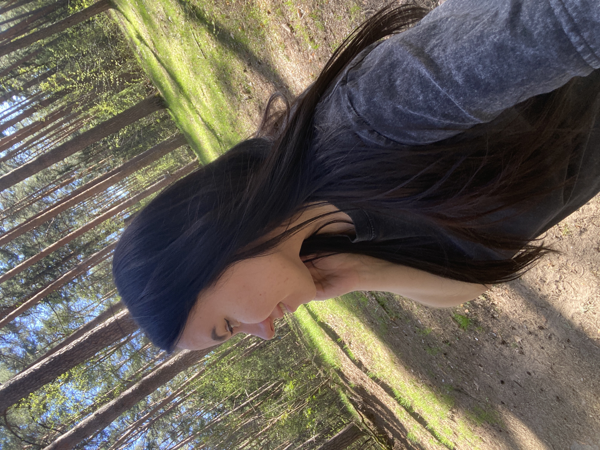 A woman with long, dark hair is standing in a wooded area. She is wearing a dark-colored, possibly grey, top. The background consists of tall trees and a path covered in dirt and some grass. The location appears to be a park or forest area within the city of Perm, Russia, indicated by the tree species and general landscape. The woman is looking down and her face is partially visible in profile.
