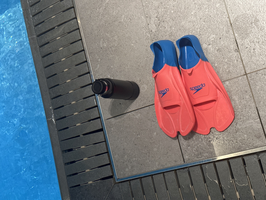 This media file presents a poolside scene, likely indoors, in London, United Kingdom. The main subjects are a pair of coral pink and blue Speedo swimming fins and a black cylindrical water bottle, all resting on grey tiled flooring.
The fins are positioned side-by-side, with the "Speedo" brand name distinctly visible in white lettering on the coral section of each fin. The blue heel straps of the fins are prominent. The black water bottle lies horizontally on the tiles, situated closer to the pool's edge, between the fins and the pool.
To the left, the bright blue water of a swimming pool is visible, showing subtle ripples on its surface. A grey slatted drainage system acts as a border between the pool and the tiled area. The tiling itself is a neutral grey, providing a clean and functional backdrop. The lighting appears to be artificial, casting soft shadows, consistent with an indoor pool environment or evening hours. The scene suggests an imminent or recently concluded swimming activity, with the equipment placed neatly at the water's edge. No individuals are present in the frame.