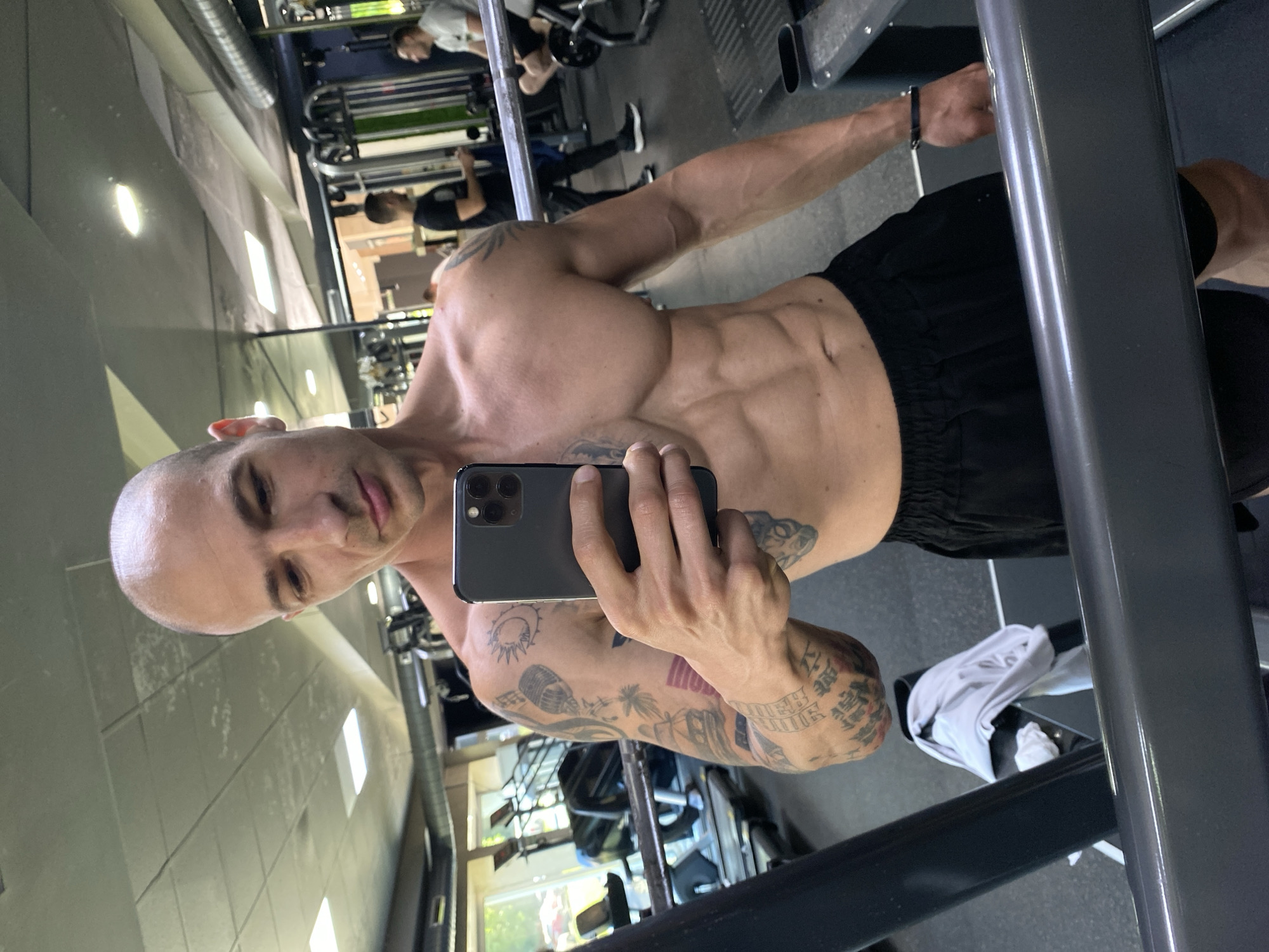 A shirtless man with a shaved head is taking a selfie in a gym. He has visible abdominal muscles and extensive tattoos on his left arm. He is holding a black smartphone, presumably to capture the image. In the background, other people are working out on various machines. The gym is located in Pleven, Bulgaria, as indicated by the photo's context. The environment includes typical gym equipment and infrastructure.