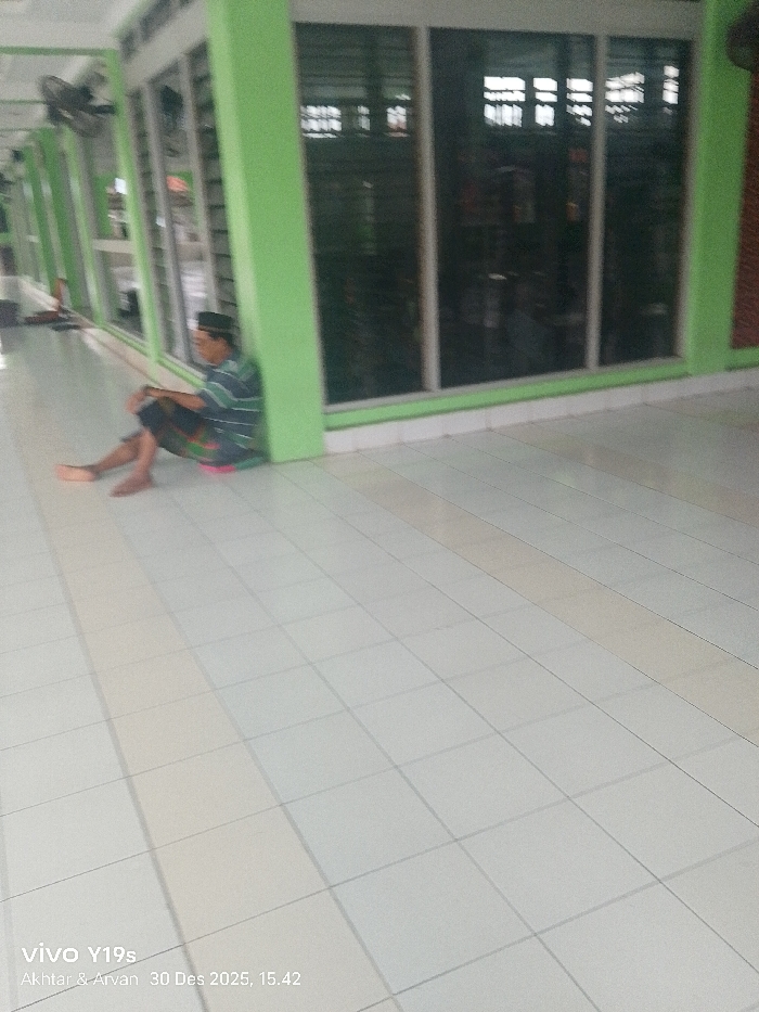 The image depicts a male individual seated on a light-colored tiled floor within a covered walkway or corridor of a building in Jagapura Kidul, Indonesia. The individual, appearing to be an adult male, is wearing a dark head covering, a striped short-sleeved shirt, and a dark patterned sarong-like garment. He is barefoot, leaning against a light green structural pillar, with a small pink object positioned beneath him. His legs are extended and slightly bent.
To the right of the seated individual, and extending further into the background, is a series of floor-to-ceiling glass window panels framed by similar light green architectural elements. The windows reflect the exterior light and reveal a dark, indistinct interior. A wall-mounted fan is visible on the upper left side of the frame. The tiled floor, light in color with dark grout lines, dominates the foreground and extends beneath the subject and toward the background. The lighting suggests daytime conditions.