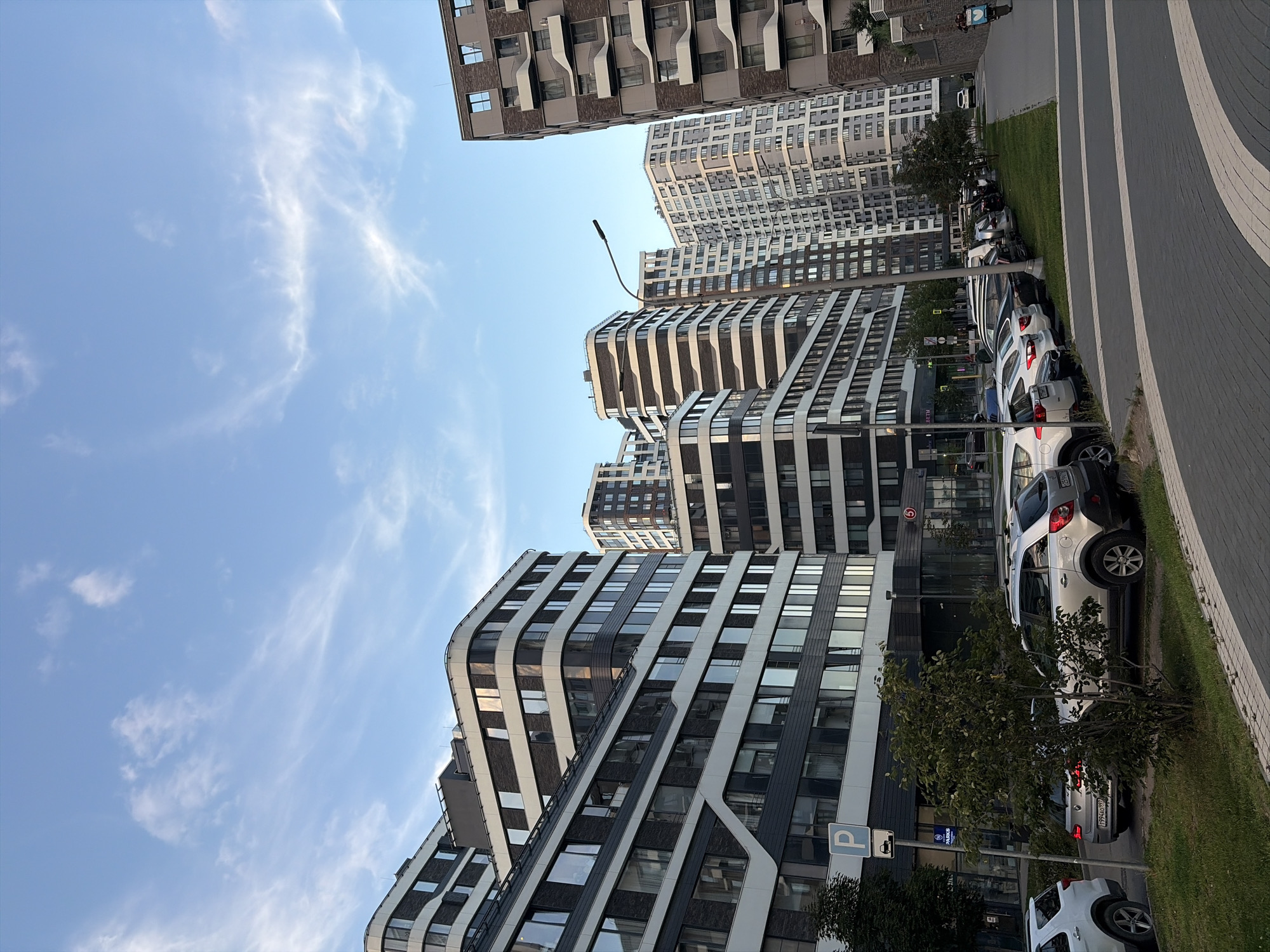 The image captures a modern urban scene in Moscow, Russia, featuring a row of contemporary residential buildings with distinctive curved facades and alternating dark and light horizontal bands. The architecture is characterized by large, reflective windows and balconies. A paved road with a pedestrian walkway is visible in the foreground, bordering a patch of green grass where several cars are parked. The sky above is a clear blue with wispy white clouds, suggesting daytime. A lamppost stands tall among the buildings, and a few trees are scattered throughout the scene. In the distance, more apartment buildings of varying heights and designs are visible, indicative of a densely populated urban development. The scene appears orderly and well-maintained, typical of a new residential district.