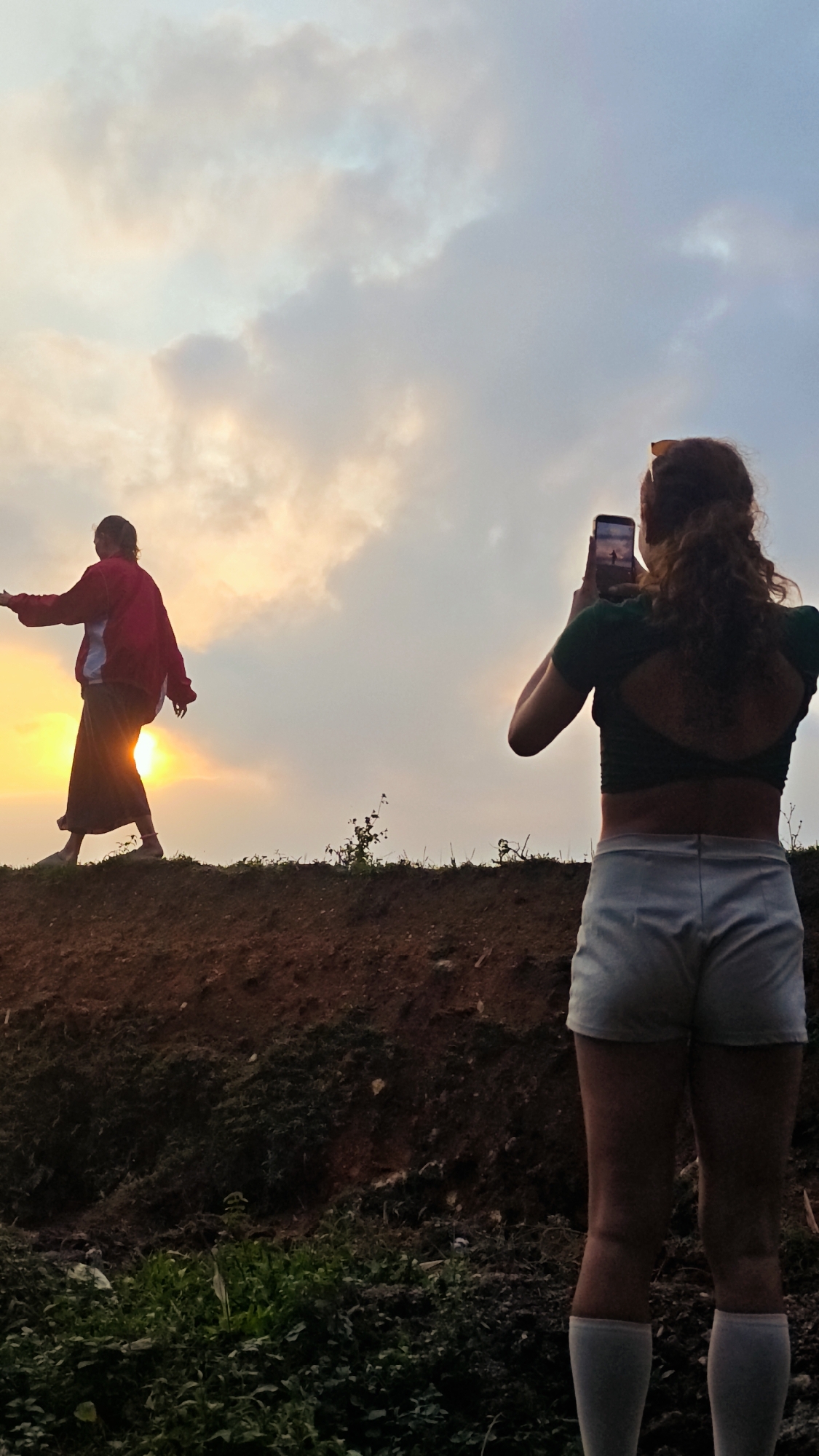 This vertical full-shot image depicts an outdoor scene during either sunrise or sunset, creating a warm, backlit atmosphere against a cloudy sky.
Two people are the primary subjects. On the left, further into the midground, a person is largely silhouetted by the sun. They wear a red jacket and what appears to be a dark, long skirt or traditional lower garment. This individual stands on a raised, uneven dirt mound, with arms slightly outstretched as if posing or balancing.
On the right, closer to the viewer and seen from behind, another person is actively taking a photograph with a smartphone held up to eye level. This person has reddish-brown curly hair pulled back, wears sunglasses on their head, a dark green crop top with a cut-out design on the back, light-colored shorts, and white knee-high socks. A faint reflection of the silhouetted person can be seen on the smartphone screen.
The setting appears to be a rural or natural environment, consistent with Nậm Dịch Commune, Vietnam. The foreground shows dark soil with scattered patches of green vegetation and small plants. The dirt mound acts as a natural barrier, separating the two individuals and contributing to the rugged landscape. The soft, diffuse light from the low sun casts long shadows and highlights the clouds, evoking a tranquil moment of capturing a scenic view. There is no visible text in the image.