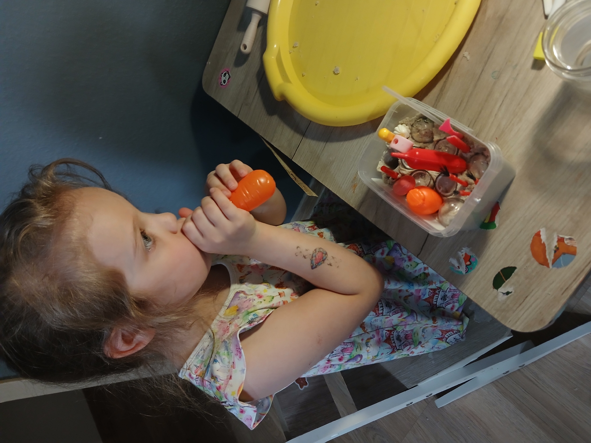 A young Caucasian girl with light brown hair, wearing a colorful dress, is sitting at a wooden table and playing with kinetic sand. She is holding an orange toy, possibly a carrot, up to her mouth. On the table in front of her is a clear plastic container filled with kinetic sand and various small toys, including red and pink plastic tools and a yellow ball. To her right, a large yellow circular tray is visible. The girl has a temporary tattoo of a gem on her upper arm. The background shows a dark blue wall and a portion of a white high chair. The flooring appears to be light wood laminate. The image is a close-up shot focusing on the girl and her activity.