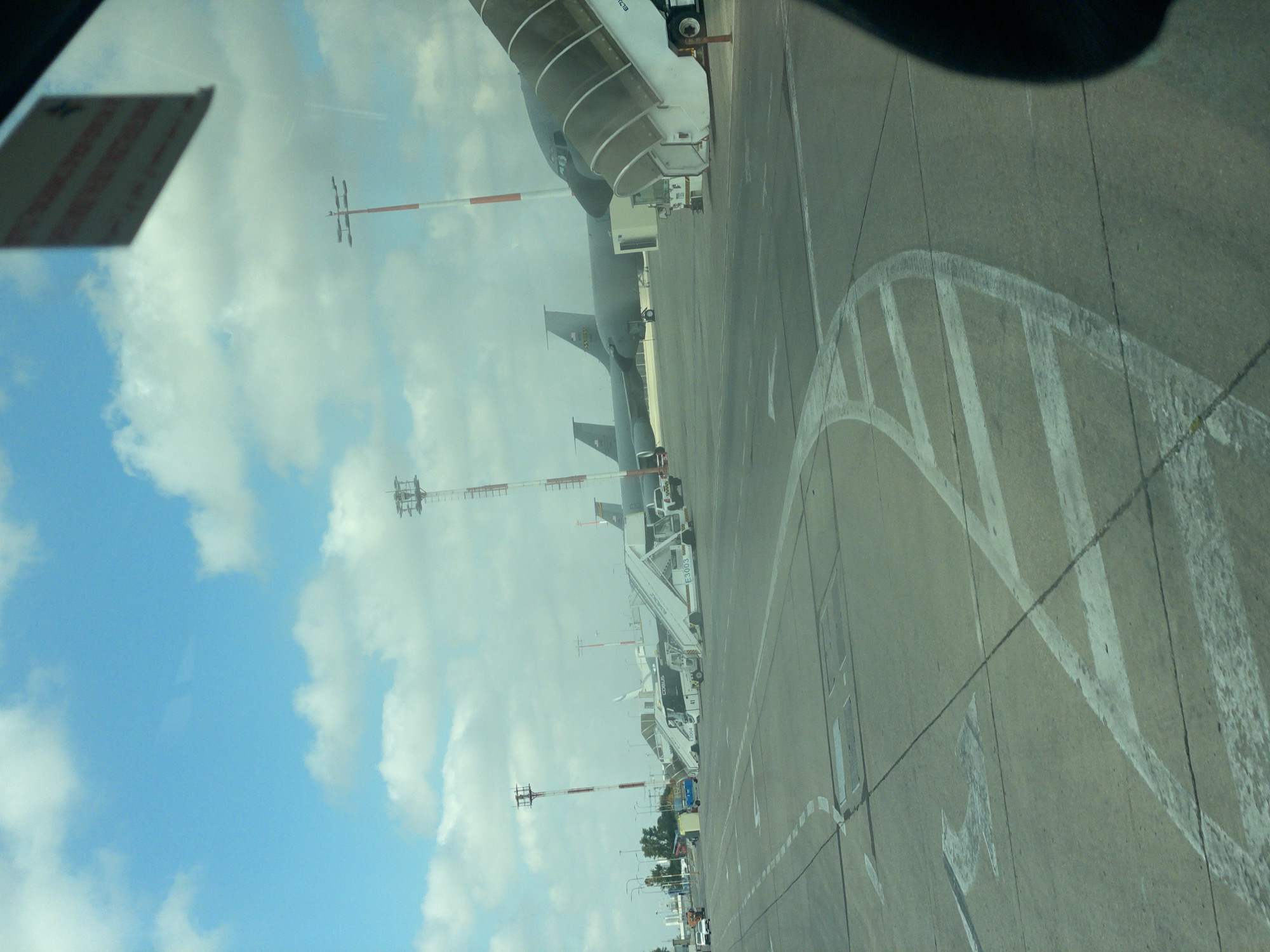 The image depicts an airport tarmac on a cloudy day, with the sky a brilliant blue with white cumulus clouds. In the foreground, the concrete surface of the tarmac is marked with white painted lines, including directional arrows and a large curved pattern. To the right, a portion of a black object, possibly a vehicle's interior or a person's head, is visible.
Further back on the tarmac, several aircraft are parked. The most prominent is a large, dark-colored jet with a distinctive hump-like structure on its fuselage, suggesting it may be a military tanker aircraft. Two other similar aircraft are parked behind it, also with their tails visible.
Adjacent to the aircraft are mobile staircases, some of which are connected to the planes, indicating passengers or crew may be boarding or disembarking. A white bus with "COBUS" written on its side is positioned near one of the staircases. Also visible are support vehicles and lights on tall poles with red and white striped markings, common airport navigational or lighting towers.
In the distance, a line of trees and some buildings can be seen, hinting at the airport's surroundings. The overall scene is one of activity at an airport, with aircraft, ground support equipment, and infrastructure present. The location is identified as Yagel, Israel, suggesting this is an airport in that region.