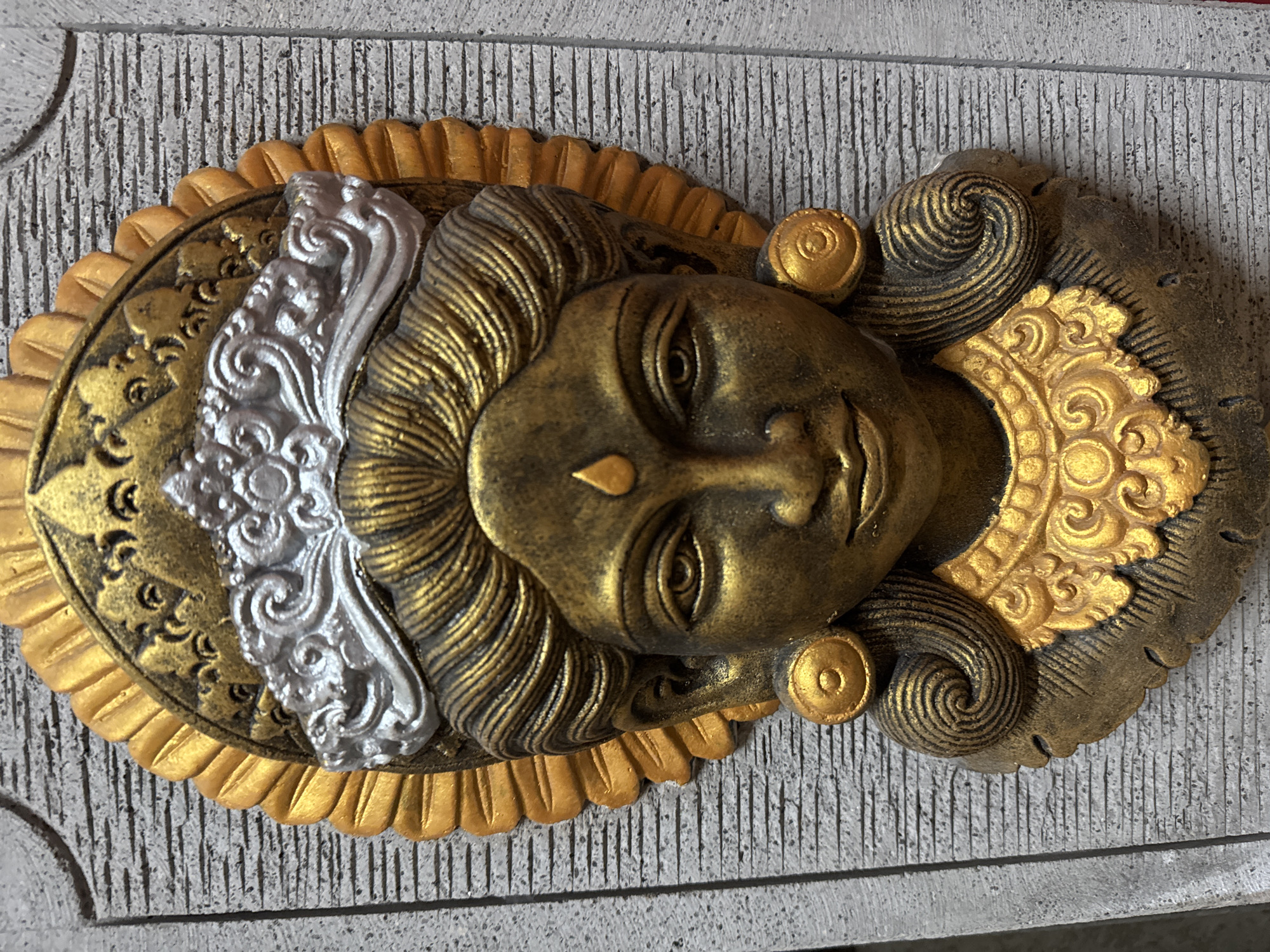 This media file is a close-up, overhead shot of a decorative wall hanging. The primary subject is a stylized, golden-brown face with intricate detailing. The face appears serene or neutral in expression. It is adorned with gold and silver elements, including a headdress or crown with decorative swirls and patterns, and what appears to be hair depicted with textured lines. The face is set against a backdrop that resembles carved stone, with a textured, vertical lined pattern. Surrounding the face is an ornate, oval-shaped decorative piece with gold accents, including a scalloped edge. The overall aesthetic suggests a cultural or religious artifact, possibly inspired by Balinese or Indonesian art, given the Kuta, Indonesia location context. There is no visible text in the image. The lighting appears to be natural and even, suggesting it was taken indoors or on an overcast day. The object is the sole focus of the image, with no other subjects or activities depicted.
