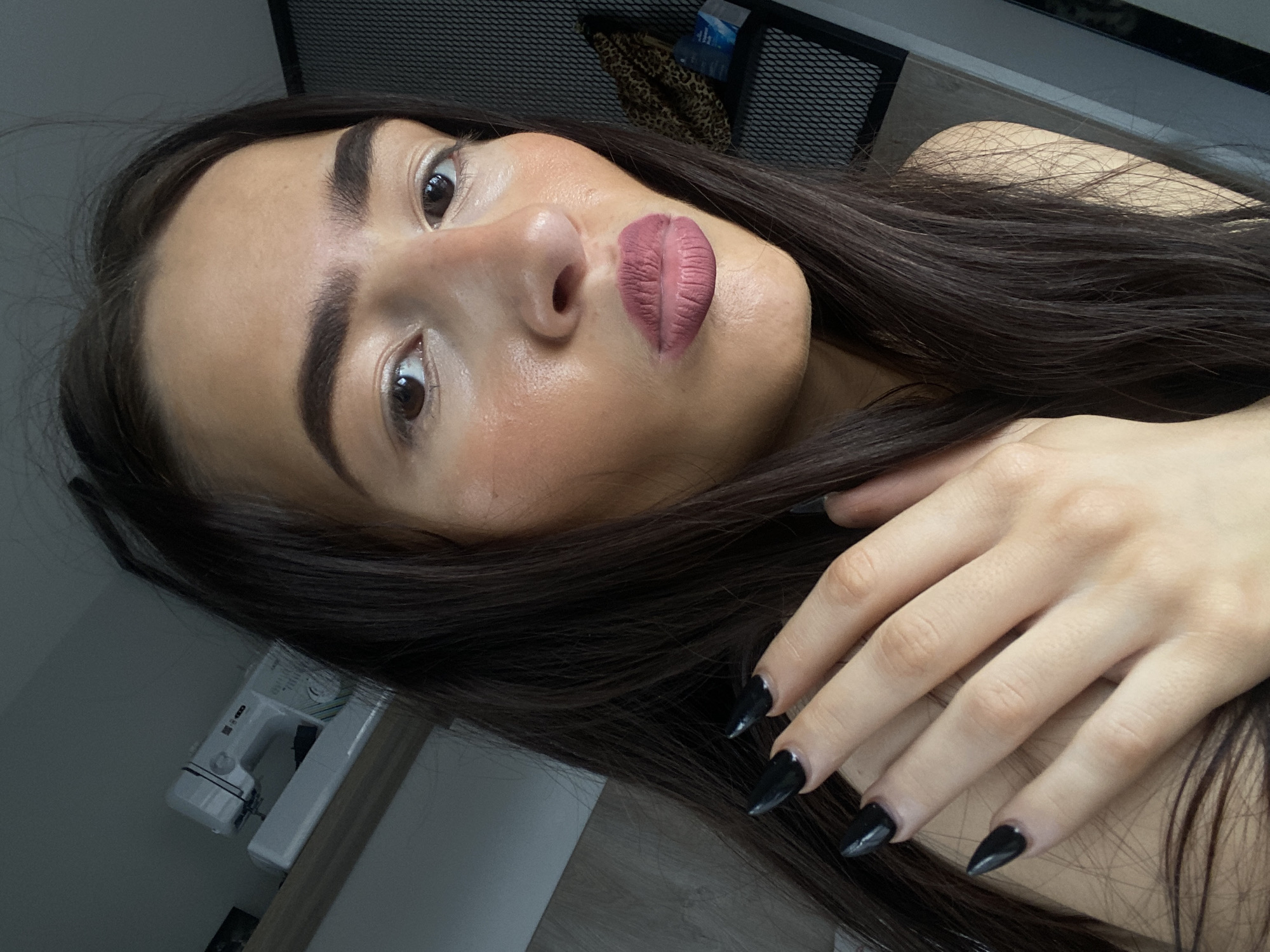 An indoor, close-up portrait in the city of London, United Kingdom, features a female subject positioned from the chest up, gazing directly at the camera with pursed lips. Her long, dark brown hair falls over her shoulders. Her right hand, adorned with long, black, pointed artificial nails, rests across her left shoulder. The subject has an olive skin tone, visible blush/contour on her cheeks, dark eyebrows, and wears dark pink matte lipstick.
The background reveals a domestic environment. To the upper left, a white sewing machine rests on a light wooden shelf. To the upper right, a dark, perforated mesh panel is visible. Below the mesh panel, on a lower shelf, there is a folded leopard-print fabric item and a white rectangular package with blue text. The lighting illuminates the subject from the front or slightly above.
