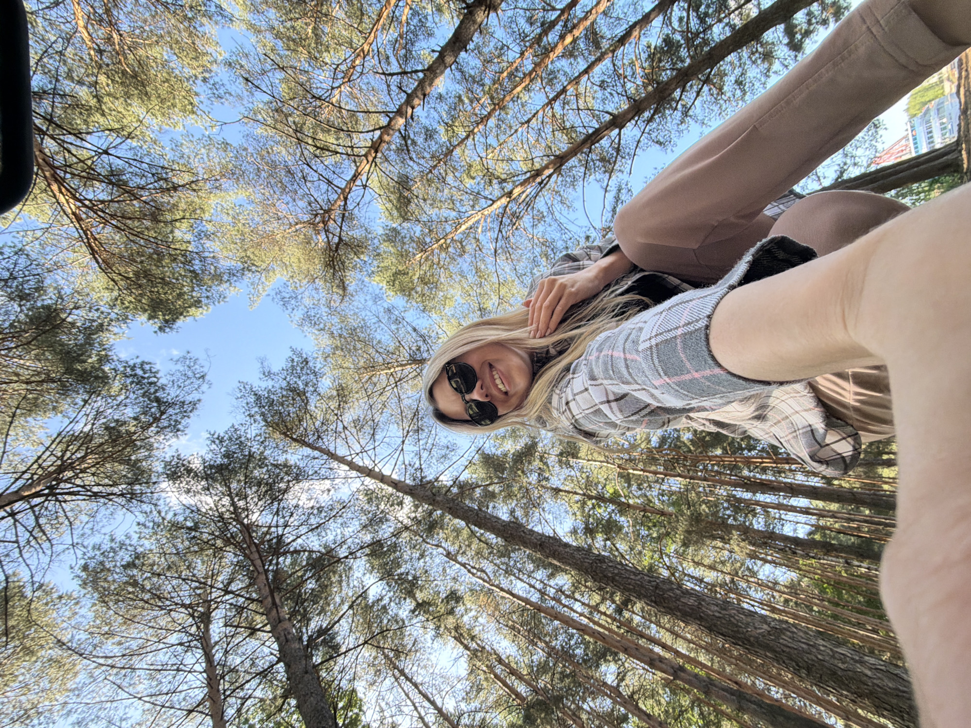 In Chaikovsky, Russia, a young woman with blonde hair and sunglasses takes a selfie from a low-angle perspective. She is wearing a plaid jacket over a dark top and tan pants. She is smiling and looking at the camera, with her hand resting on her chest. The background consists of tall pine trees reaching towards a clear blue sky. Some distant buildings can be seen through the trees.
