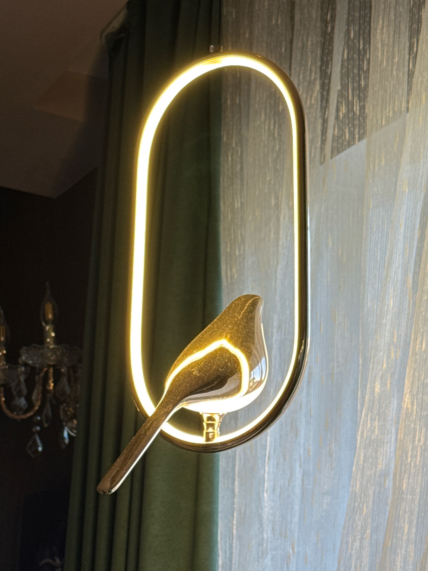 The image shows a close-up of a modern pendant light fixture shaped like an oval frame with an illuminated inner edge. A stylized, metallic bird sculpture is perched within the frame, its body catching the light. The background consists of draped curtains, one a dark green and the other a sheer, light-colored fabric, suggesting an indoor setting, possibly a room with windows. To the left, a portion of a more traditional chandelier with multiple arms and crystal-like elements is visible. The lighting is warm, emanating from the pendant and ambient sources. There are no people or visible signs of the city of Torzhok, Russia in the image.