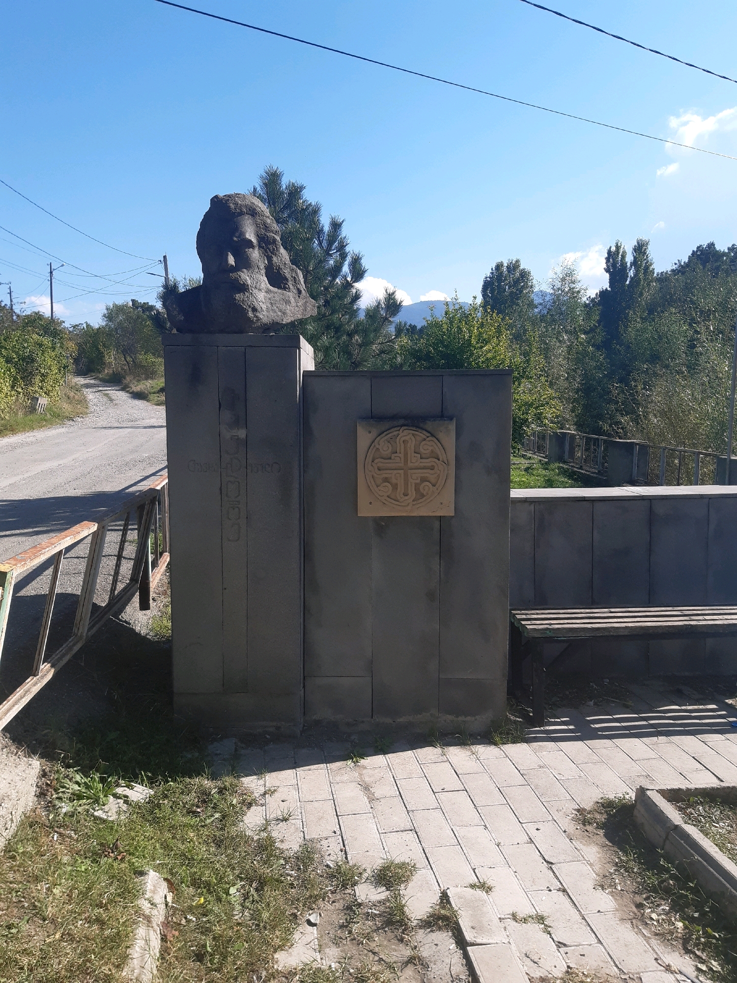In Garikula, Georgia, a grey stone bust of a man with a beard and long hair stands atop a grey stone monument. To the right of the bust, a square stone plaque with a circular cross design is mounted on the monument. To the right of the monument, a wooden bench sits on a paved walkway. The monument is situated outdoors, with a road and trees visible in the background. The sky is clear and blue.