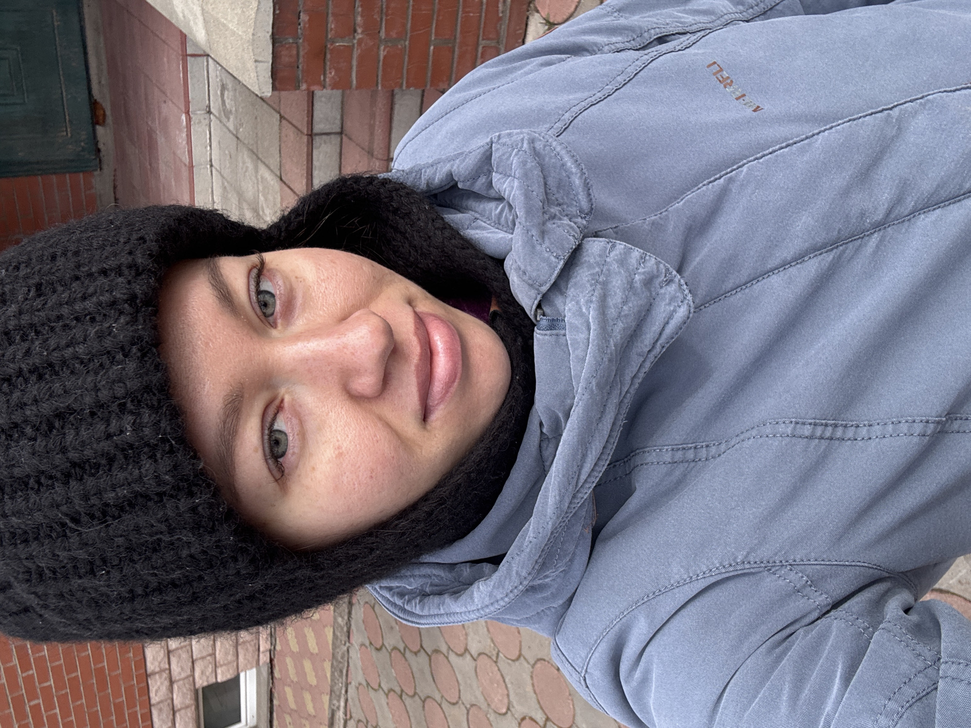 A woman in a black knit hat and a light blue jacket is posing for a selfie outdoors. Her face is tilted towards the camera. The jacket collar is visible. The background consists of a red brick wall, and a sidewalk with a pattern of beige circles. A small window is also visible in the background. The location is Ферма, Russia.