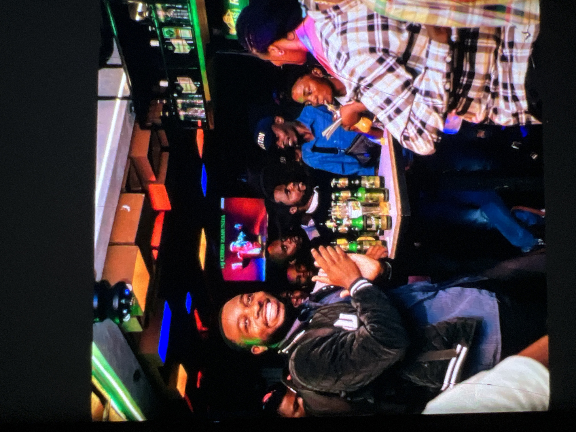 The image depicts a lively indoor scene, likely a bar or club, with a group of people gathered. The lighting is dim and colorful, with prominent neon green, blue, and red accents illuminating the ceiling and shelves. A screen in the background displays a figure in red, identified by text as "DJ CHRIS ZAMUNDA". Several bottles of alcoholic beverages are visible on shelves behind the bar. In the foreground, a Black man with a wide smile and clapping hands is the central focus, wearing a dark jacket with white stripes. To his right, other individuals are engaged in conversation and drinking. A woman in a plaid shirt is also prominent in the right foreground, holding a drink. The overall atmosphere suggests a social gathering or event. While the image is from Lagos, Nigeria, specific details about the exact location within the city are not discernible from the visual information.
