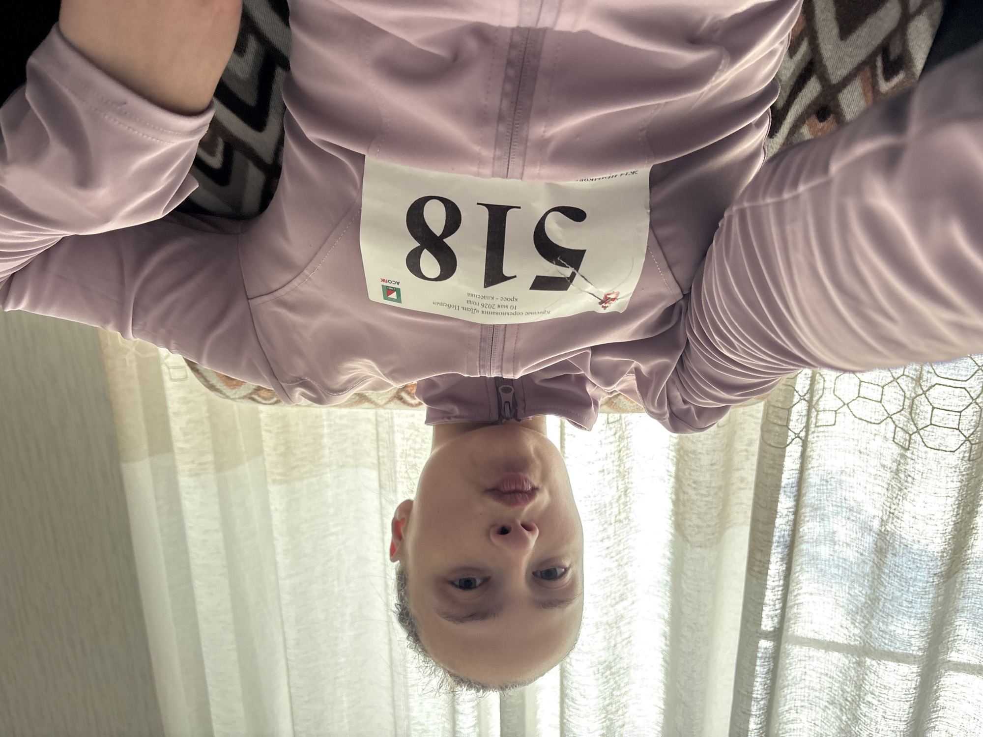 A person with a race bib (number 518) is upside down in a photograph. The bib has the logo for ASOPIK and information in Russian about the "Day of Victory" competition, a cross-country classic race held on May 10, 2026. The person is wearing a pinkish-purple athletic jacket. Behind the person is a window with white curtains and a couch with a patterned cover. There is no explicit information in the image to confirm it is taken in Perm, Russia, only that the text is in Russian.