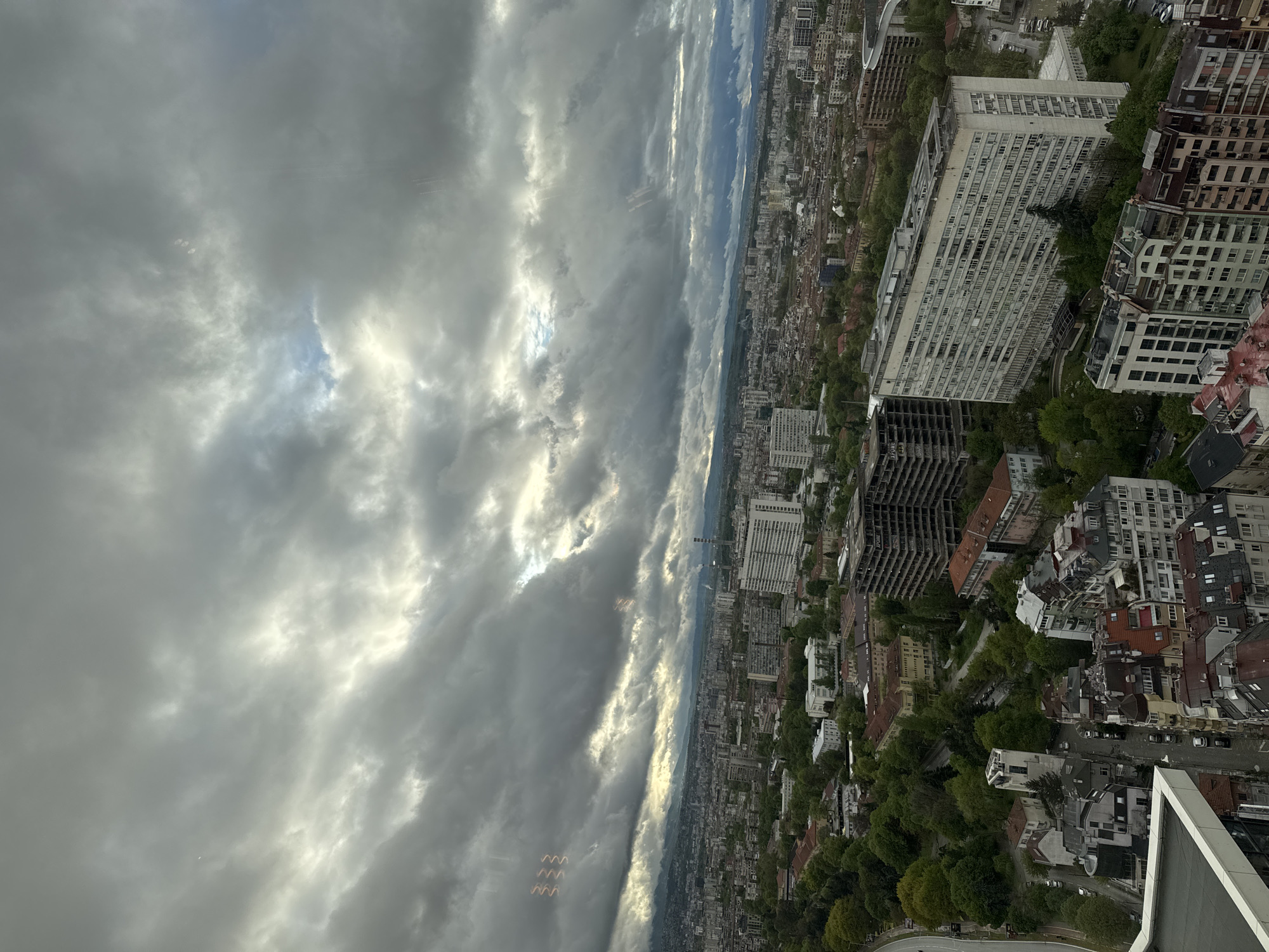 This high-angle, panoramic image captures a vast urban landscape, likely Sofia, Bulgaria, viewed from a significant height. The city stretches to the distant horizon under a dramatic, cloudy sky.
The scene features a dense mix of buildings, including prominent large, multi-story white blocks and numerous other residential and commercial structures of varying architectural styles. In the mid-ground, a long, white building with many windows stands out, alongside the skeletal remains of an unfinished or partially demolished building with exposed rebar. Extensive greenery, including trees and planted areas, is interspersed throughout the urban environment, softening the built-up areas.
The sky is largely covered with thick, dark gray clouds, but brilliant patches of sunlight break through, creating dynamic light rays and highlights. This suggests either early morning or late afternoon lighting conditions. Notable details include several light-colored, zigzag or wavy patterns in the upper left quadrant of the image (as rotated), which appear to be reflections on a window or lens artifacts. No people or specific activities are discernible, providing a static, expansive view of the cityscape. There is no visible text in the image.