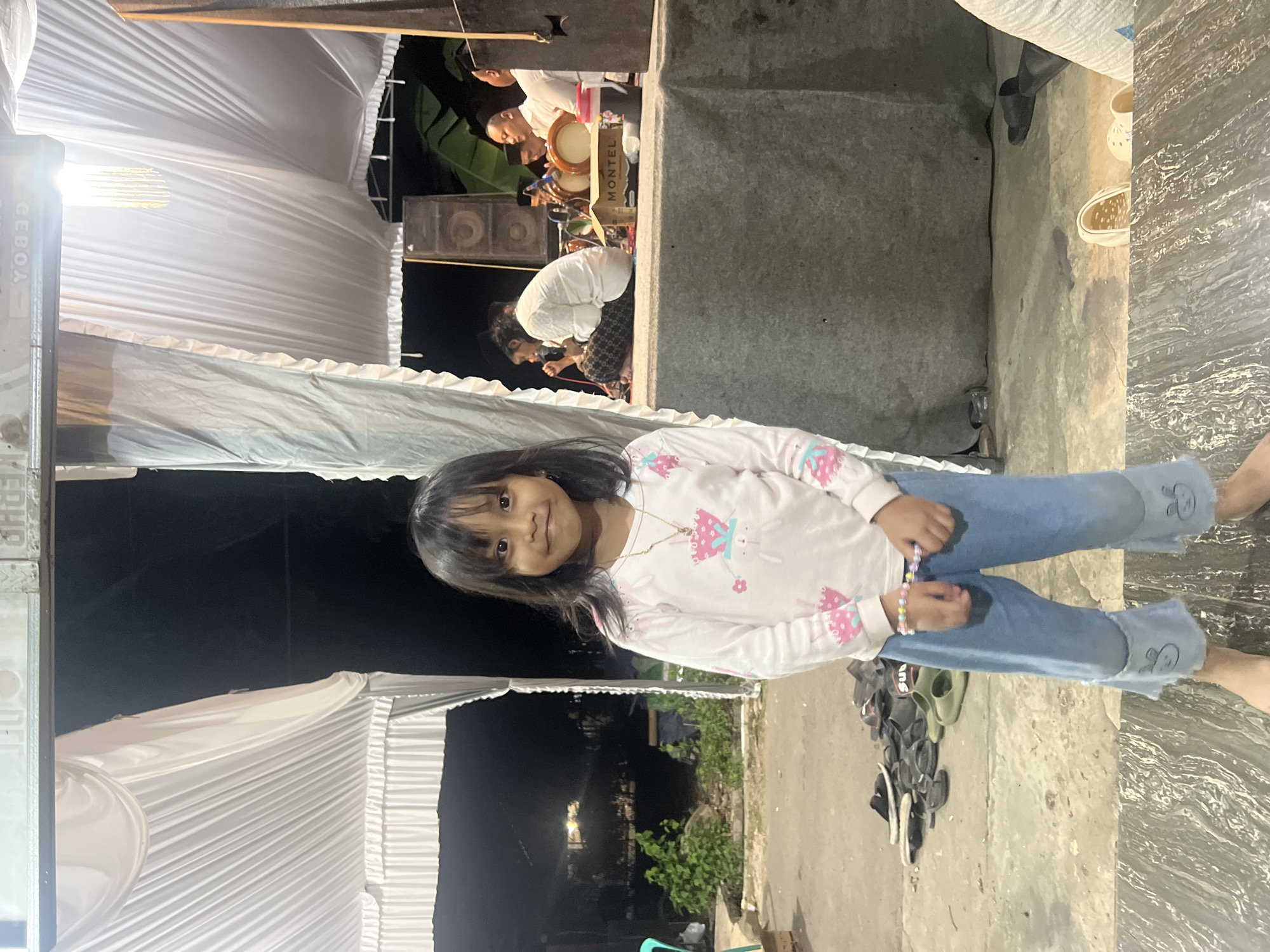 A young girl, appearing to be of Indonesian descent and dressed in a long-sleeved pink and white shirt with cartoon graphics and blue jeans, stands in the foreground of an outdoor scene. She is facing the camera with a slight smile, her hands clasped in front of her. Behind her, a group of men are seated on a raised platform, some of whom appear to be playing musical instruments, possibly drums. The area is decorated with white draping fabric, and there are speakers visible. The ground is a concrete surface with several pairs of sandals scattered about. The background suggests a rural or semi-rural setting with some vegetation and structures visible in the dim light. The overall ambiance suggests an informal gathering or performance, possibly a cultural event, taking place in the evening. The image is taken in Wedarijaksa, Indonesia, a sub-district in the Pati Regency of Central Java.