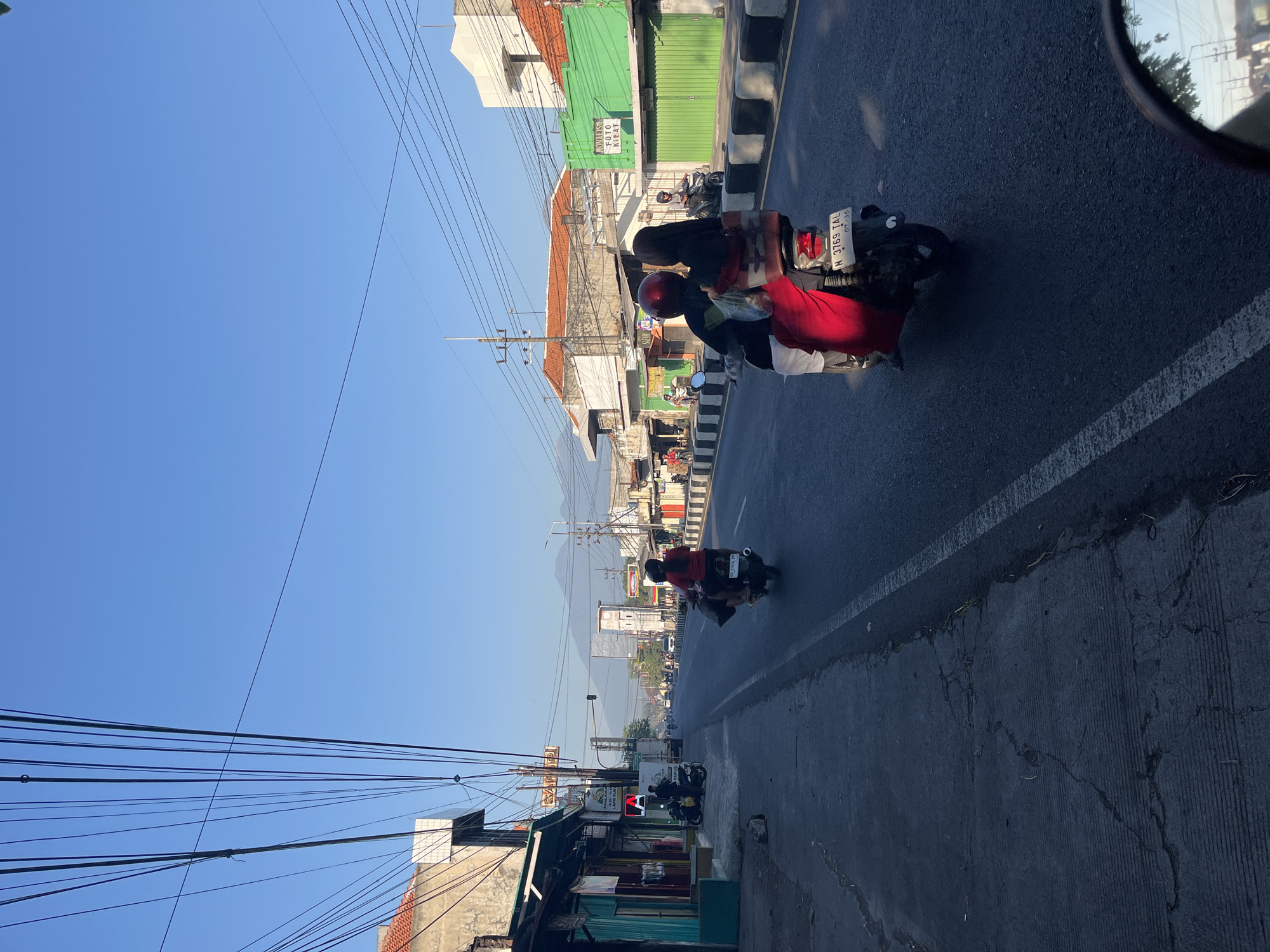 This image depicts a street scene in Porong, Indonesia, on a clear day with a bright blue sky. The foreground shows the asphalt road with a white lane divider. In the middle ground, two people are riding a scooter, with the passenger wearing a red garment and a black headscarf. The scooter's license plate reads "H 3769 TAL". On the left side of the road, there are shops and buildings, including one with a turquoise facade and another with a sign that appears to be an advertisement. On the right side of the road, there are more buildings, some with green storefronts, and a sidewalk lined with black and white concrete bollards. In the distance, more buildings and possibly mountains are visible under the clear sky. Overhead, a dense network of power lines crisscrosses the scene. The overall atmosphere is that of a typical bustling street in an Indonesian city.