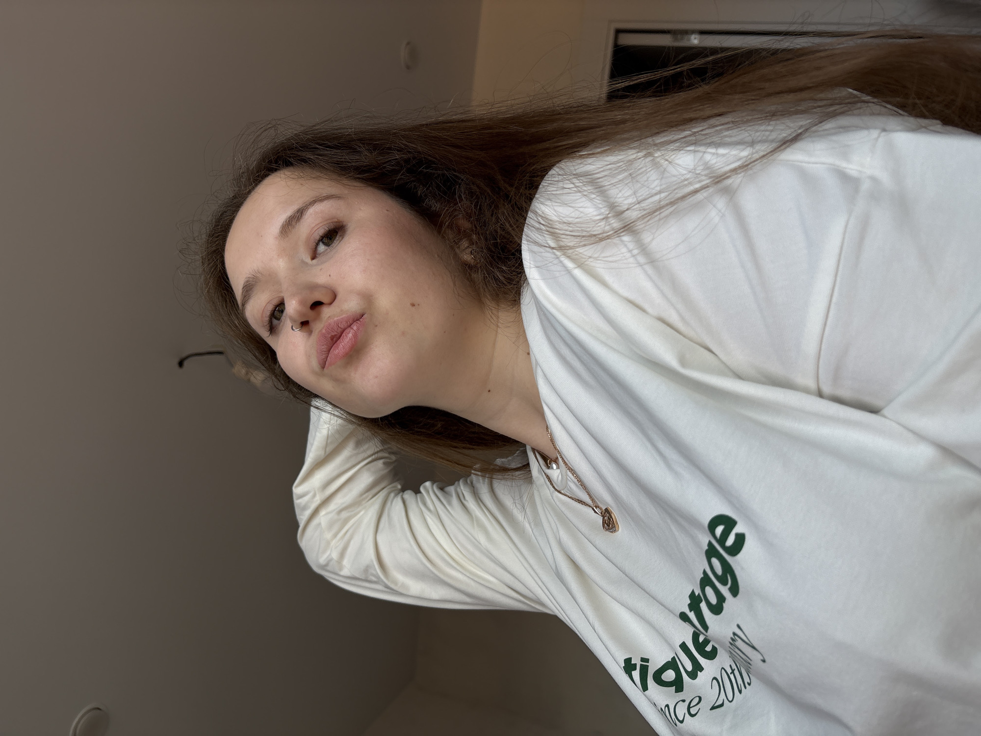 A woman with long brown hair and a nose ring is looking at the camera. She is wearing a white long-sleeved shirt with green text. The background is a light-colored wall and ceiling, with a glimpse of a dark object, possibly a window frame, in the upper right corner. No information regarding Izhevsk, Russia is discernible in the provided image.
