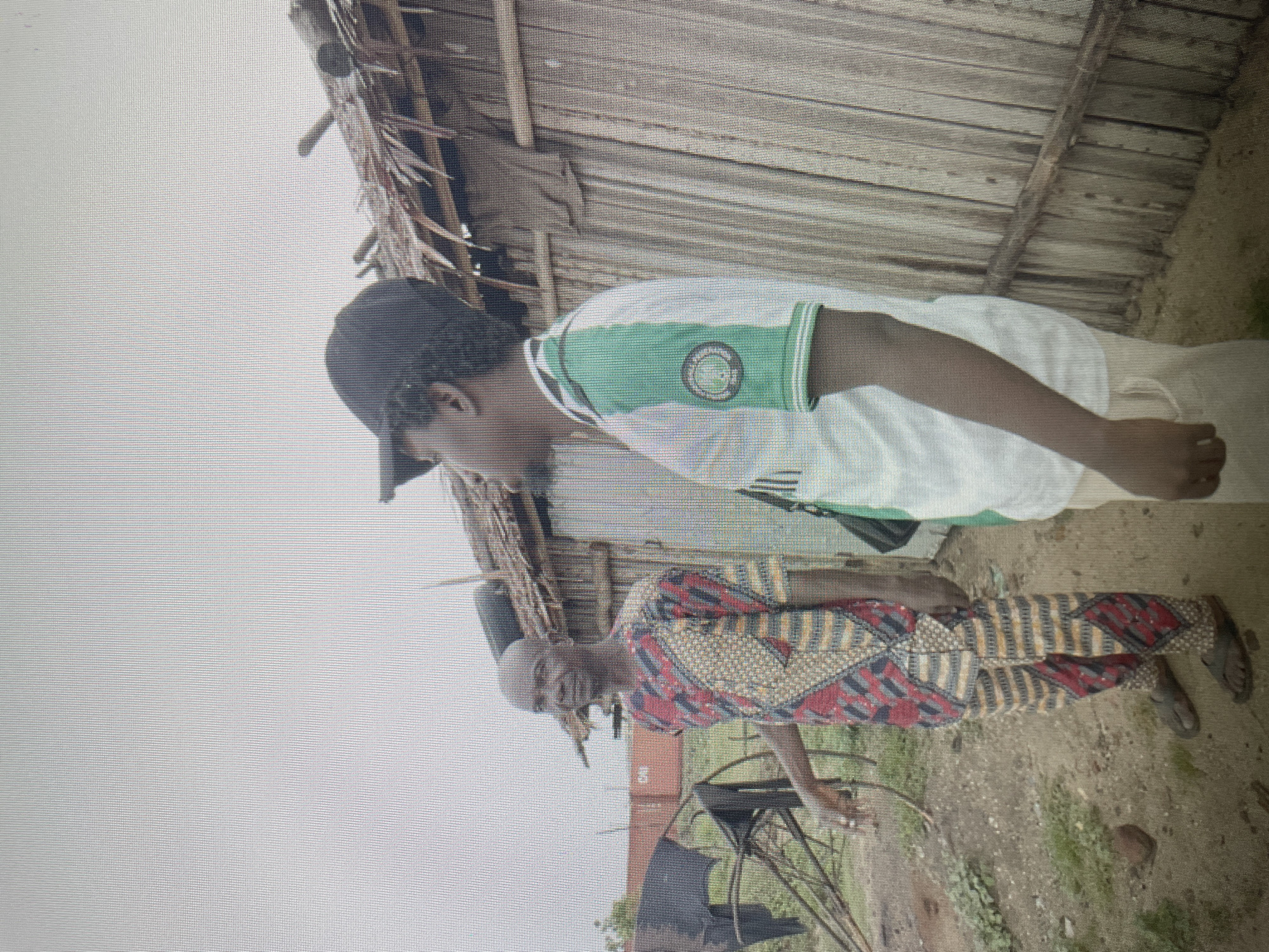 A medium shot shows two males standing outside a simple structure with a thatched roof and wooden plank walls in an unnamed city. The younger man is wearing a white shirt with green trim and a Nigerian soccer federation logo, a black baseball cap, and tan pants. The older man is wearing a patterned red, gold, and blue tunic and pants. They are standing on what appears to be dirt and sparse ground cover. In the background are some metal structures and indistinct greenery. The sky is overcast.