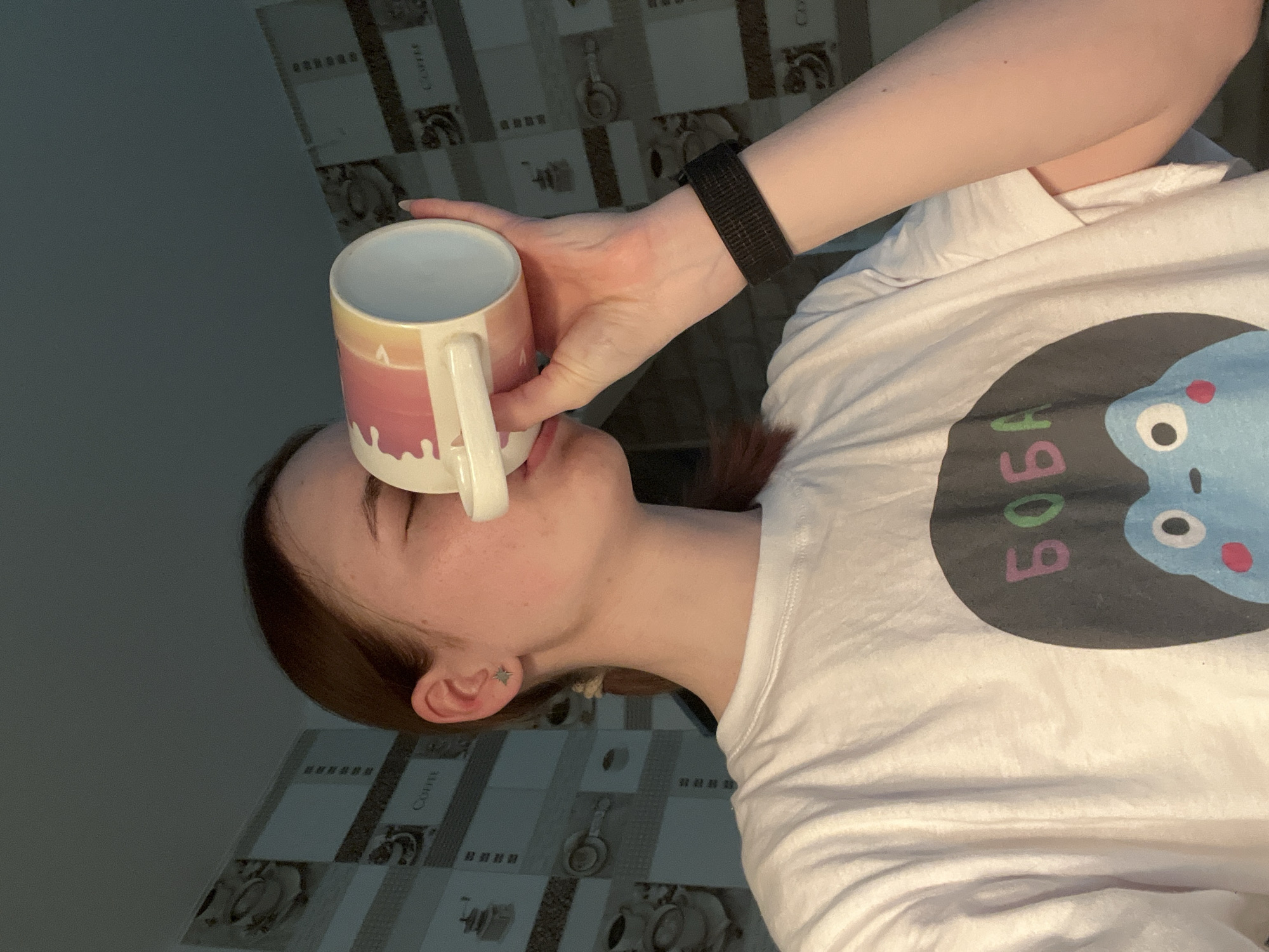 A young woman is holding a ceramic mug with a handle up to her forehead. The mug has a white interior and a gradient exterior of yellow, orange, and pink, with white paint dripping effect along the bottom. She is wearing a white t-shirt with a cartoon character print that has the word "BOBA" printed on it. She has brown hair and is wearing small star-shaped earrings. On her wrist, she wears a black elastic wristband. The background features a tiled surface with a pattern of kitchenware images, indicating a kitchen setting. The photograph appears to be taken indoors. It is not possible to determine the location in the city of Chaikovsky, Russia.