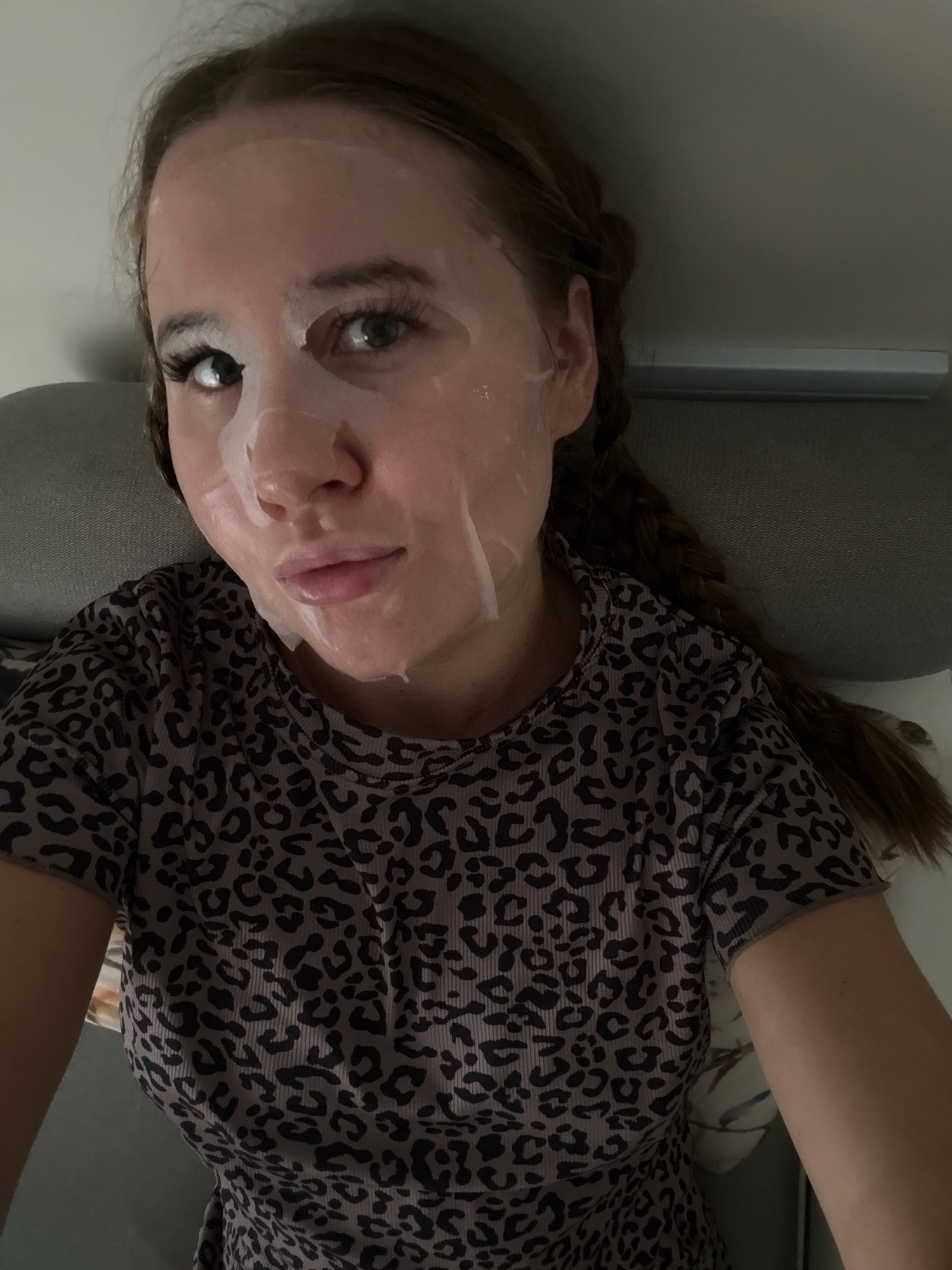 A young Caucasian female with light skin and brown hair is shown from the chest up. She is wearing a gray shirt with a black leopard print and has a sheet mask on her face. Her hair is styled in two braids. She is lying down, with her head resting on a gray pillow. The background is a plain wall. The scene appears to be indoors, possibly a bedroom. There are no visible landmarks or indicators to suggest the location is Yiwu, China.
