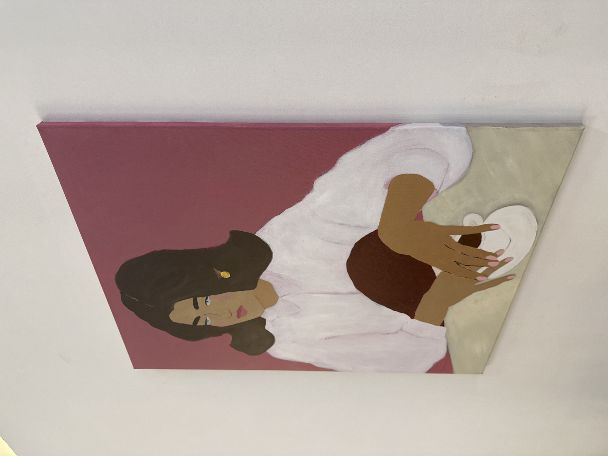 This is an image of a painting depicting a person reclining on a surface. The painting is oriented with the top of the image on the left. The subject has dark hair and is wearing a white garment with voluminous sleeves. They are holding a white object, possibly a bowl or a plate, with both hands. The hands have light skin with pink fingernails. The face of the subject is visible, with light skin, blue eyes, and pink lips. A small, round, gold stud is visible in the hair. The background of the painting is divided into two distinct color fields: a deep pink on the left and a pale beige on the right. The painting is displayed on a wall with a white ceiling visible above. The context indicates the location as Moscow, Russia, but there are no specific visual cues in the painting to confirm this. There is no visible text in the image.