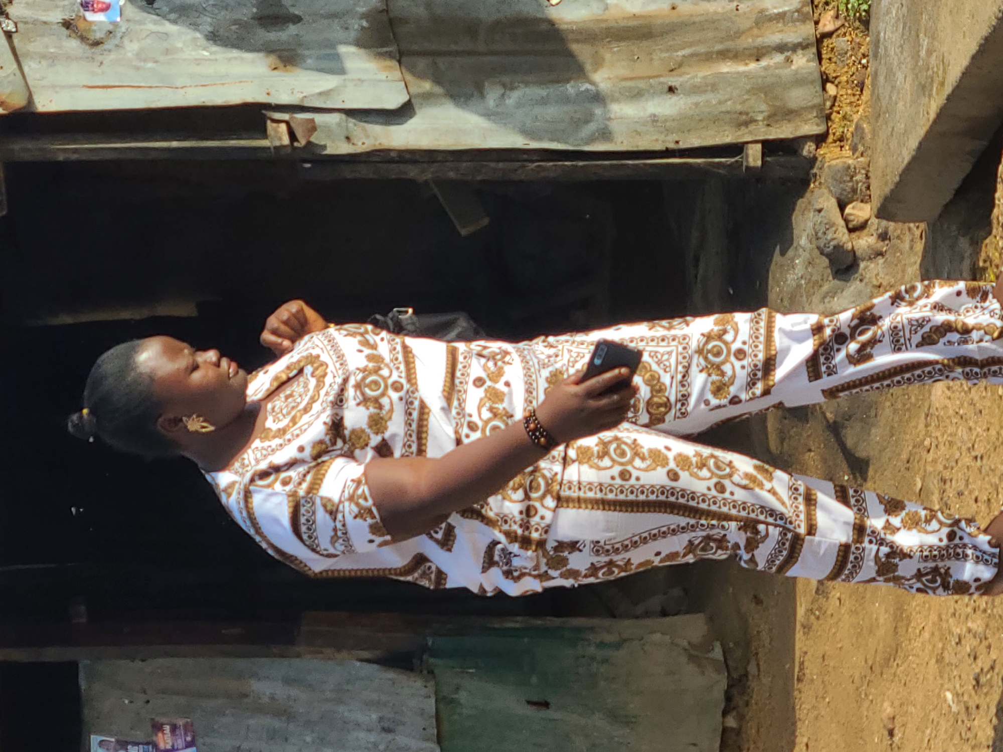 A person, identified as a woman, is captured in a vertical orientation, holding a smartphone in her right hand. She is wearing a white outfit adorned with a repeating pattern of ornate, golden motifs. The outfit consists of a long-sleeved shirt and matching trousers. Her hair is pulled back, and she has a distinct, gold-colored earring in her left ear, shaped like a leaf or feather. A beaded bracelet is visible on her right wrist.
The background suggests an outdoor, somewhat rustic setting. Behind the woman, there is a dark, open doorway or recess, possibly leading into a building or structure. Above this opening, there are corrugated metal sheets, appearing weathered and aged. To the right of the woman, the ground is covered in dry, sandy soil, with some rocks and sparse vegetation visible. A concrete structure, possibly a wall or ledge, is also present to the right. The lighting indicates it is daytime, with sunlight casting shadows, suggesting it might be a sunny day. The overall impression is that of an everyday scene in an outdoor environment, possibly in a residential or market area. No specific text is clearly legible in the image.
