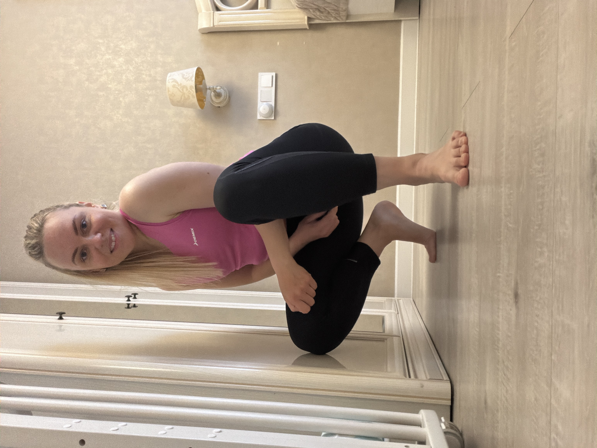 An indoor portrait-oriented image, rotated 90 degrees counter-clockwise, features a young woman smiling brightly at the camera while performing a complex yoga or stretching pose. She has long, straight blonde hair and a cheerful expression, looking directly into the lens.
The woman is wearing a vibrant pink athletic tank top, with the brand name "DEMIX" visible in white text on her left chest, and black leggings. She is barefoot. Her body is in a deep, seated twist; her left leg is bent with the foot tucked near her right hip, and her right leg is bent and crossed over the left. Her arms are wrapped around her legs in a bind, showcasing her flexibility.
The background shows the interior of a room, likely in a home. The wall is a light beige or cream color with a subtle textured finish. A white, ornate wall sconce with a cream-colored shade is mounted high on the wall, alongside a white light switch and dimmer control. Below these, a white door or cabinet with decorative paneling and small black handles is visible. The floor, appearing on the right side of the rotated image, is a light wood-patterned laminate or tile. Part of a white radiator or drying rack is visible in the lower foreground. The lighting suggests it is daytime. The image captures a moment of physical activity and happiness, set in an indoor environment, potentially in Votkinsk, Russia.