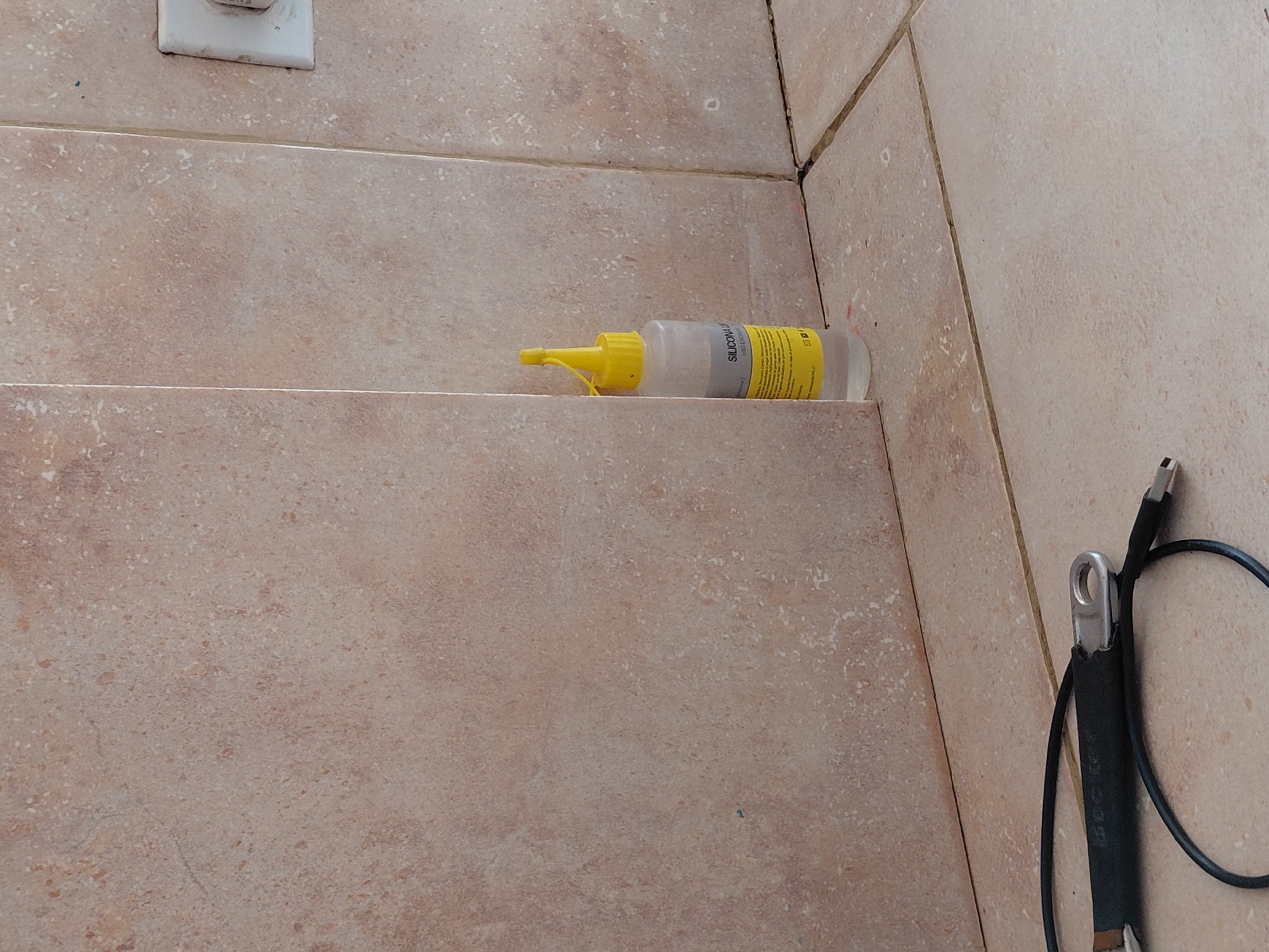 The image captures a close-up, low-angle view of a tiled indoor or semi-enclosed space in Santa Ana, Venezuela. The scene features light brown or beige-colored tiles with a speckled, stone-like texture, used on both vertical walls and at least one horizontal surface, possibly a shelf or a step. Grout lines, some appearing dark, define the edges of the tiles and the corner where two walls meet.
Prominently placed on a horizontal tiled surface is a clear plastic squeeze bottle with a yellow cap and nozzle. The label on the bottle is partially visible, clearly showing the word "SILICONA," indicating it contains silicone sealant or adhesive. Below the silicone bottle, near the right edge of the frame, a black electrical cable is visible, coiled loosely on a lower tiled surface. One end of the cable features a silver metallic lug or ring terminal, while the other end appears to have a different connector, possibly a USB plug. In the upper left corner, a portion of a white electrical outlet or switch cover is attached to the tiled wall.
There is no activity taking place, and no people are visible. The objects are static, suggesting a pause in work or items left in a utility area. The lighting is even, without strong shadows, making the time of day indeterminate.