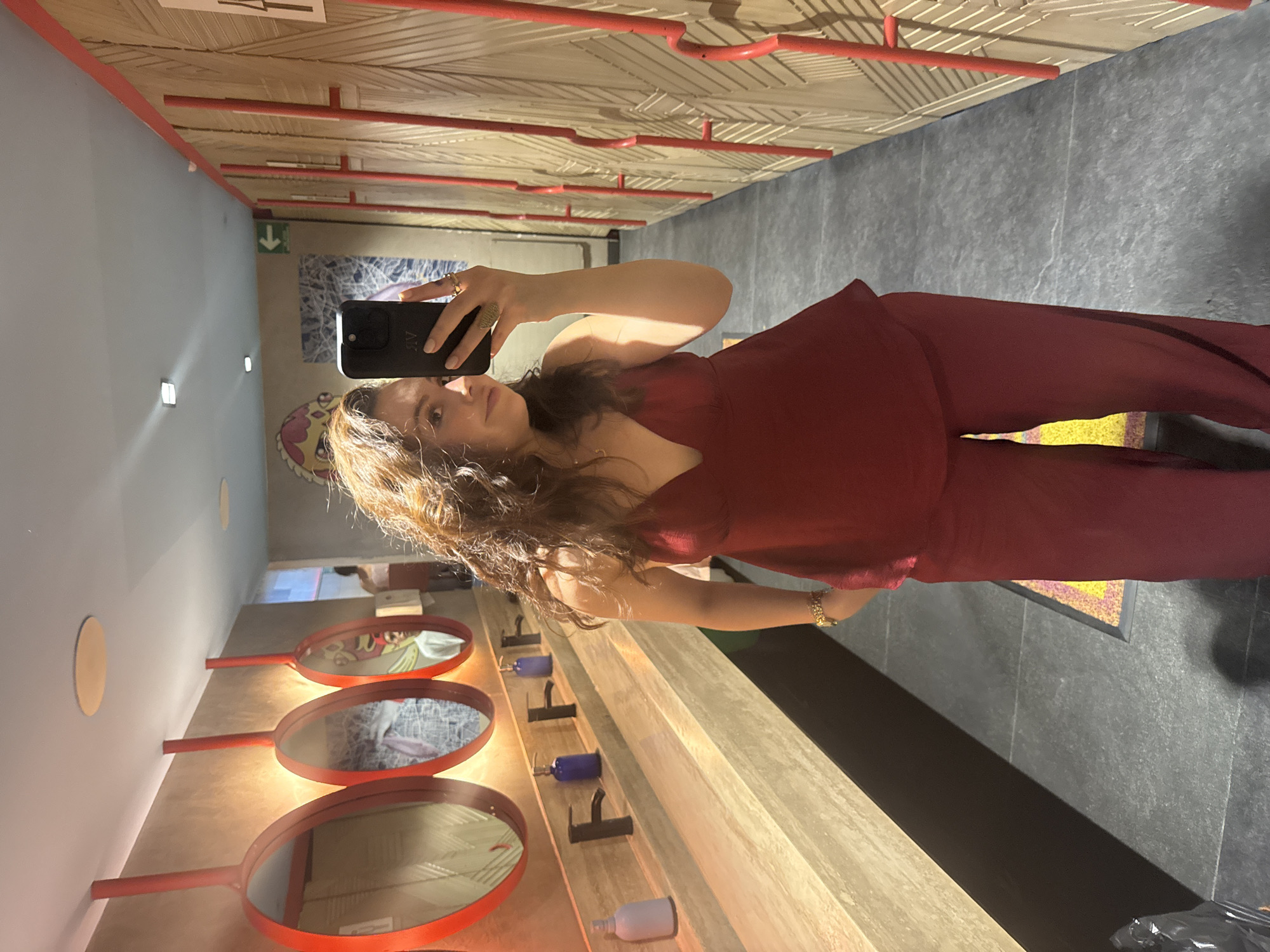 A woman is taking a selfie in a brightly lit room. She's holding a black smartphone with the camera facing her. She has long, brown hair and is wearing a red pantsuit. The room contains several red-rimmed mirrors resembling hand mirrors mounted on a wall. Below the mirrors are bottles of various colors attached to the wall. The floor is gray tile. The background includes a red-piped ceiling and a sign with a green arrow pointing towards an exit. The city is Mexico City, Mexico.