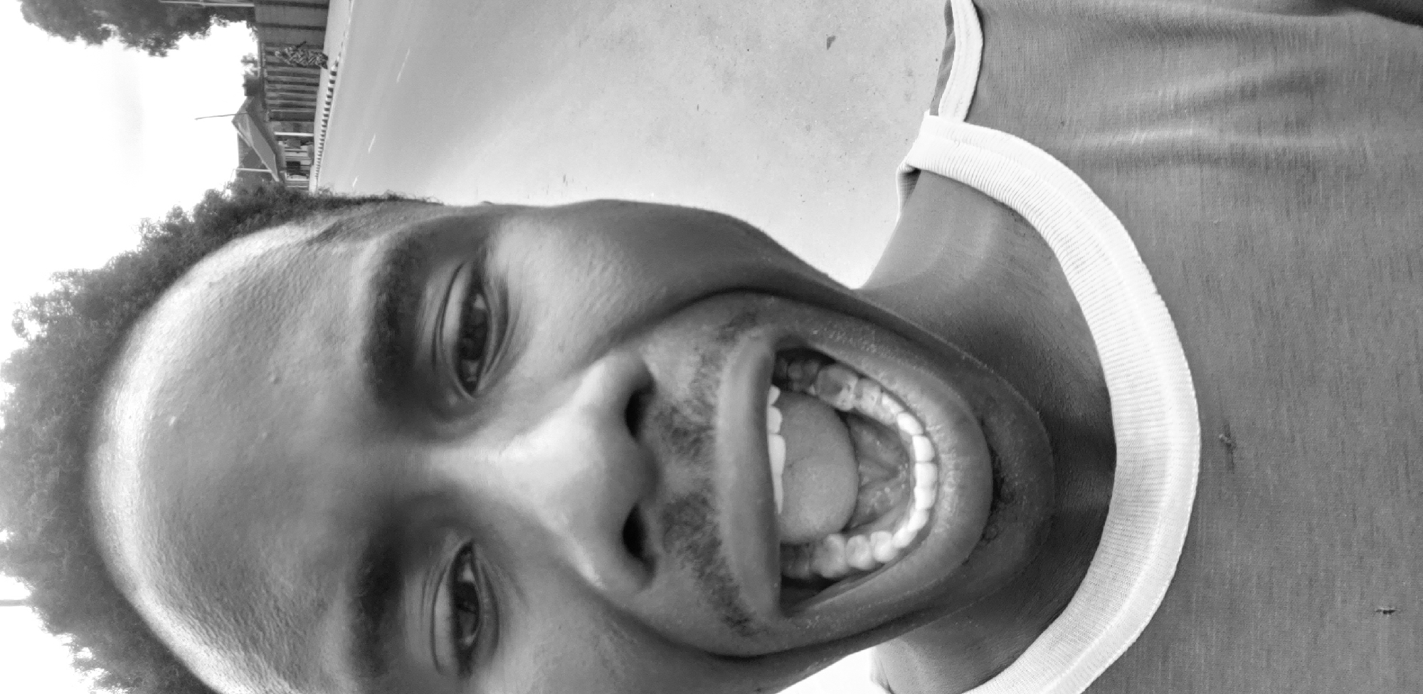 This is a black and white, close-up, upside-down selfie of a young Black man. He is wearing a white-trimmed shirt, possibly a tank top. The man has dark, curly hair and a light mustache. His mouth is wide open, revealing his teeth and tongue. His eyes are open, and he appears to be smiling or laughing. The background suggests an outdoor setting, possibly a street in Narayi, Nigeria. The lighting indicates it is daytime, and the weather appears to be overcast. The image is slightly grainy, typical of older or lower-resolution photography. There is no visible text in the image.