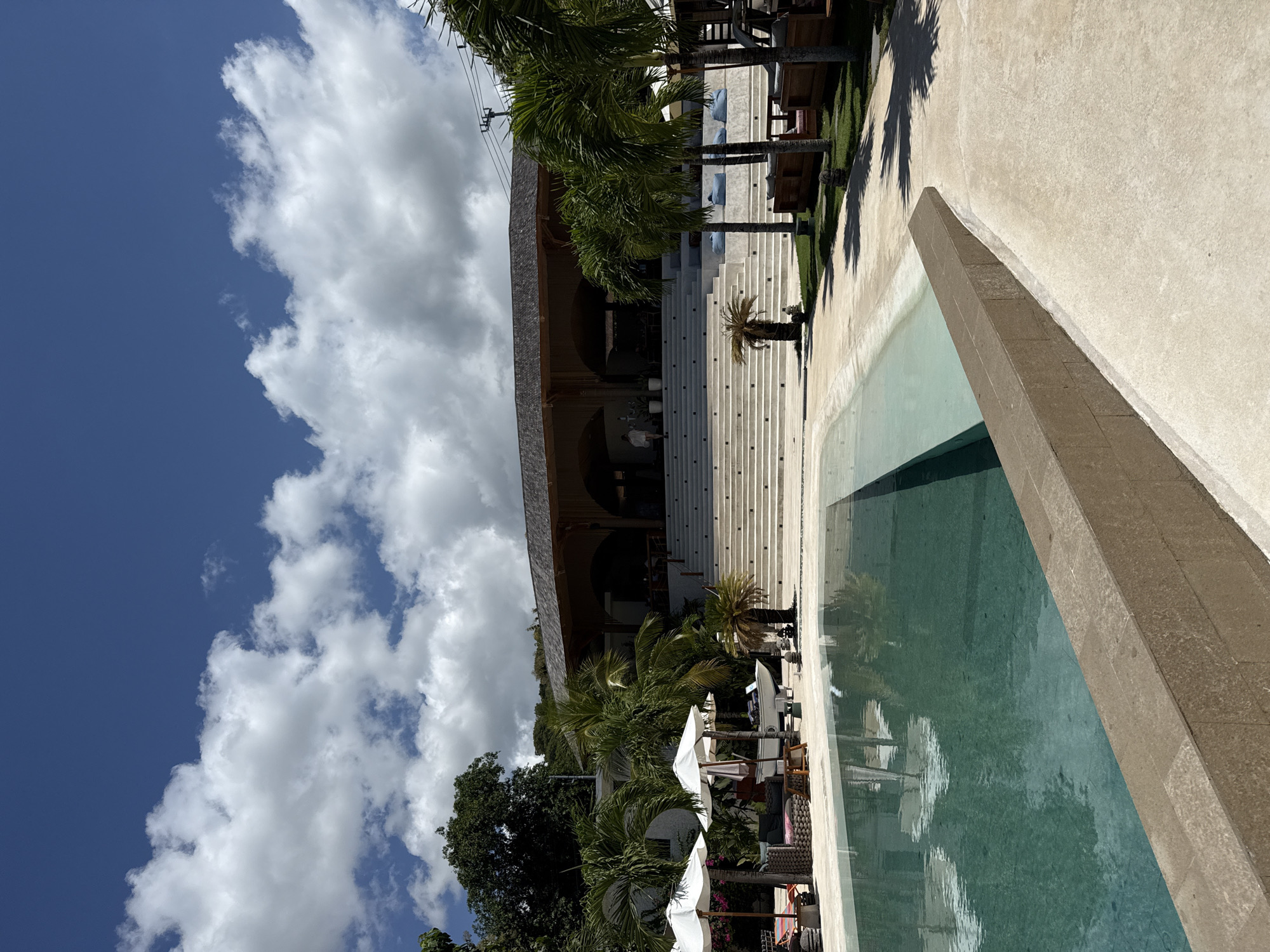 The image shows a poolside scene at a resort or villa in Nusa Penida, Indonesia. A long, rectangular swimming pool with greenish water occupies the right side of the frame. The pool's edge is made of light brown stone. Parallel to the pool is a wide, light-colored concrete walkway. Beyond the walkway, palm trees line the edge of a building with a large staircase leading up to an arched entrance. The staircase is light gray. A few small palm trees are positioned along the base of the stairs. White parasols, lounge chairs, and tables are set up near the pool, suggesting a relaxation area. The sky is blue with large, white cumulus clouds. The entire scene is well-lit, indicating a sunny day.