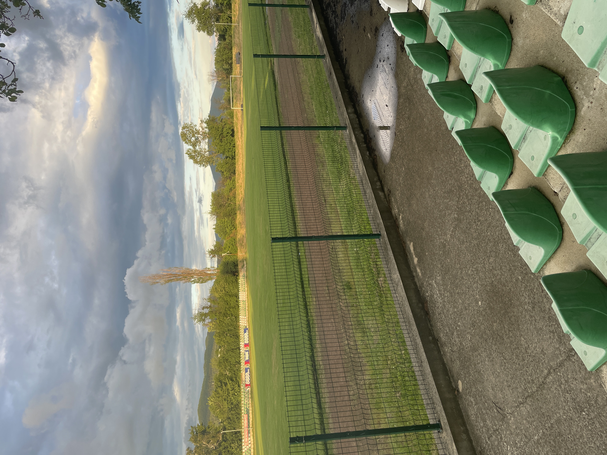 The image, shown in a vertical orientation but depicting a horizontal scene, features an empty football (soccer) field and stadium seating under a dramatic, cloudy sky, likely in Ihtiman, Bulgaria.
In the right foreground, rows of bright green plastic stadium seats are arranged on concrete tiers. The concrete ground in front of the seats is wet, with visible puddles reflecting the light. A dark green chain-link fence separates this seating area from the football pitch.
The field itself is a vibrant green turf, though strips of darker, possibly worn or dirt, ground run parallel to the fence. A white goalpost is visible on the far side of the field. Beyond the pitch, a line of dense green trees and vegetation stretches towards distant mountains in the background.
The sky is largely covered with soft grey clouds, but patches of golden sunlight break through, illuminating the cloud edges, suggesting late afternoon or early evening. The wet ground indicates recent precipitation. The scene is devoid of people, creating a quiet and contemplative atmosphere. On the far side of the field, several advertising banners are visible, one prominently displaying "8888.bg" in white text on a red background, with other "8888.bg" logos also present.