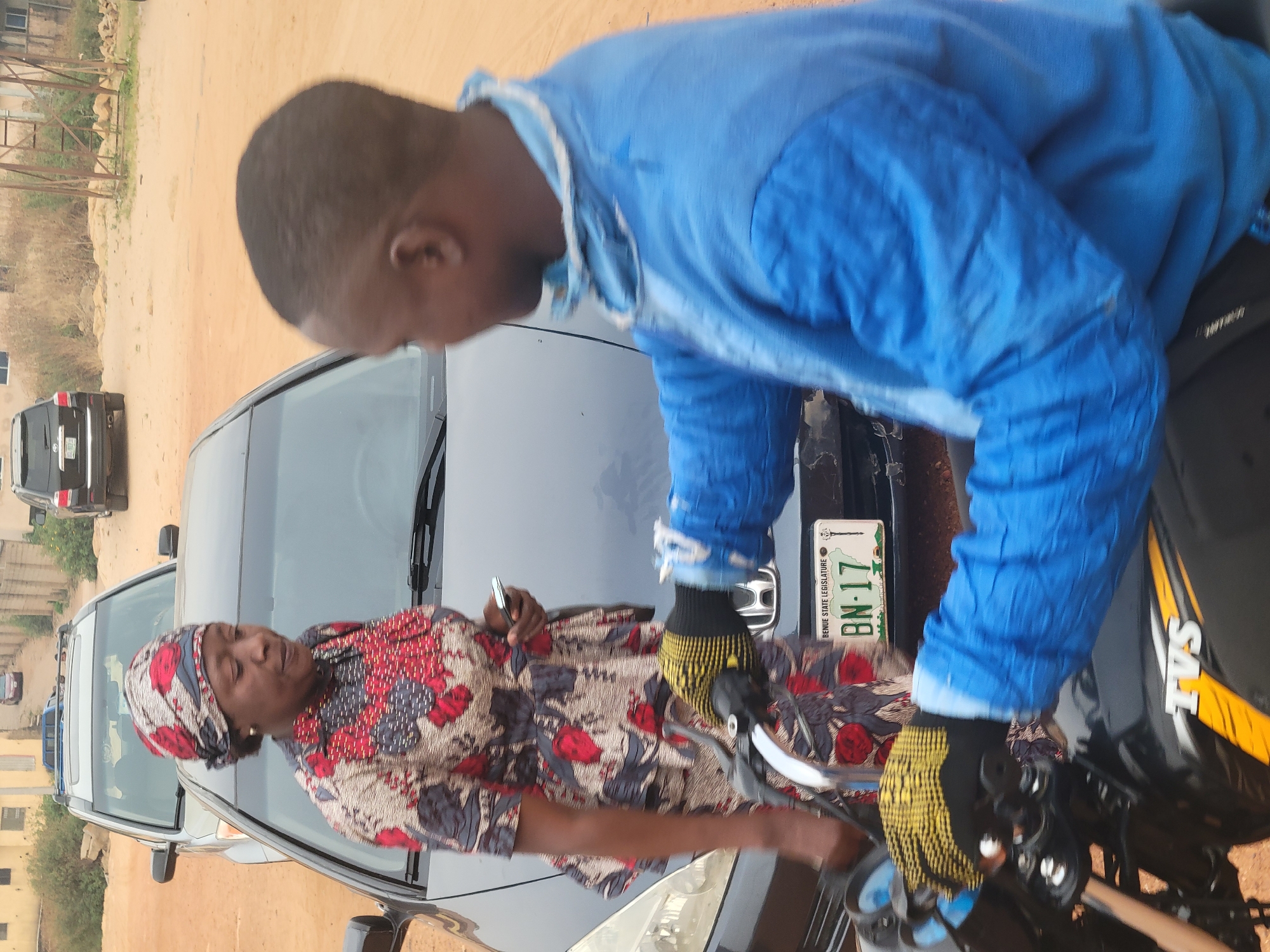 The image captures two individuals interacting near a parked black Honda car and a black and yellow TVS motorcycle in Gura Top, Nigeria. A woman with a patterned headscarf and top is seen holding a metallic object, possibly a pen or tool, in her right hand. She is standing near the front of the car, partially obscured by the vehicle. A man in a blue shirt and yellow and black gloves is leaning over the motorcycle. The car has a green license plate, partially visible, indicating "Benue State Legislature" with the inscription "BN-17". In the background, there are buildings, fences, and other parked vehicles. The ground is mostly bare earth with sparse vegetation.