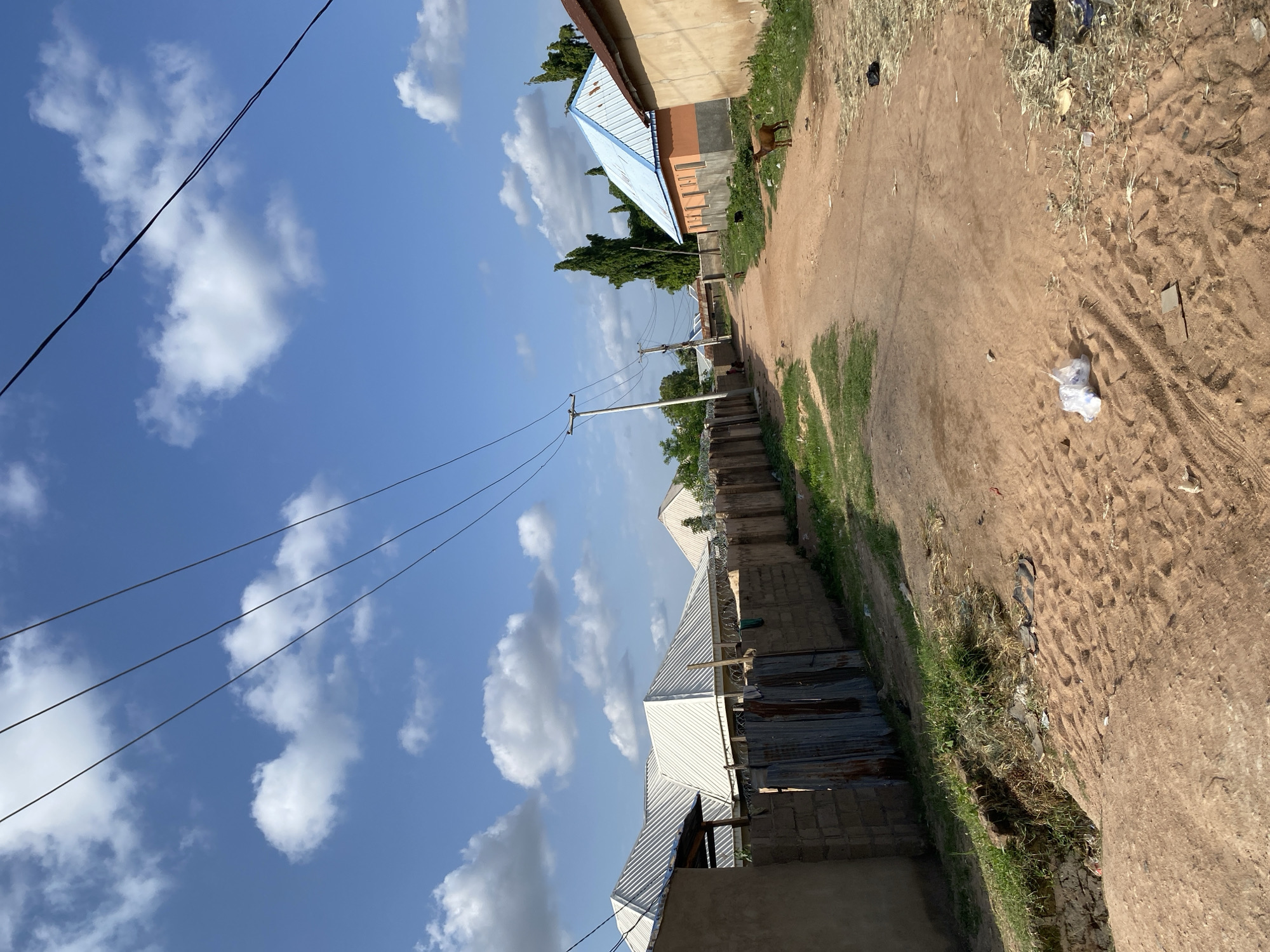 The image depicts a rural scene in Jauro Yinu, Nigeria. The setting is a dirt road with houses lining one side. The sky is a bright blue with fluffy white clouds, suggesting it is daytime and the weather is fair.
The subjects include several buildings, some appearing to be houses with corrugated metal roofs, and one with a light blue roof. There are also utility poles with visible electrical wires crossing the sky. On the side of the road, there is some sparse grass and dry vegetation, and evidence of litter, including plastic bags and other debris. In the distance, a goat is visible standing on the right side of the road.
No specific activity or event is clearly taking place. The scene appears to be a static view of a residential area. The overall impression is one of a sunny day in a somewhat unkempt but inhabited rural environment. There is no visible text in the image.