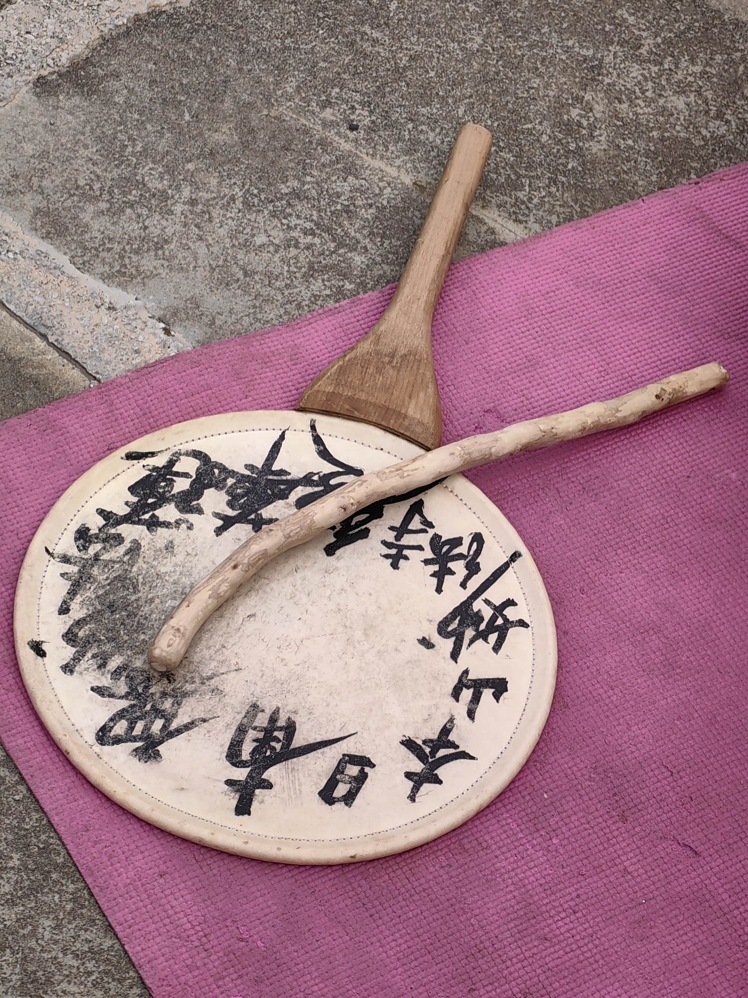 This is a close-up, high-angle shot of two wooden implements resting on a textured, vibrant pink mat. The mat is placed on a grey, speckled concrete surface.
Dominating the center of the frame is a round, off-white drumhead, likely made of animal hide, stretched over a shallow frame. The drumhead is adorned with black ink calligraphy, seemingly in a Japanese or Chinese script. A thin, light-colored, gnarled stick is positioned diagonally across the drumhead, its curve following the curvature of the drum. To the upper right of the drumhead, a flat, wooden paddle-like object with a rounded end is placed, its handle extending upwards and out of the frame. The wood of both implements appears weathered and aged.
The scene is likely located outdoors, possibly in a courtyard or public space. The concrete appears to be paving stones or a similar surface. The overall impression is one of artistic or cultural items laid out for display or use.
There are no people visible in the image. The image's context in Vashisht, India, might suggest a connection to local crafts, spirituality, or performance.